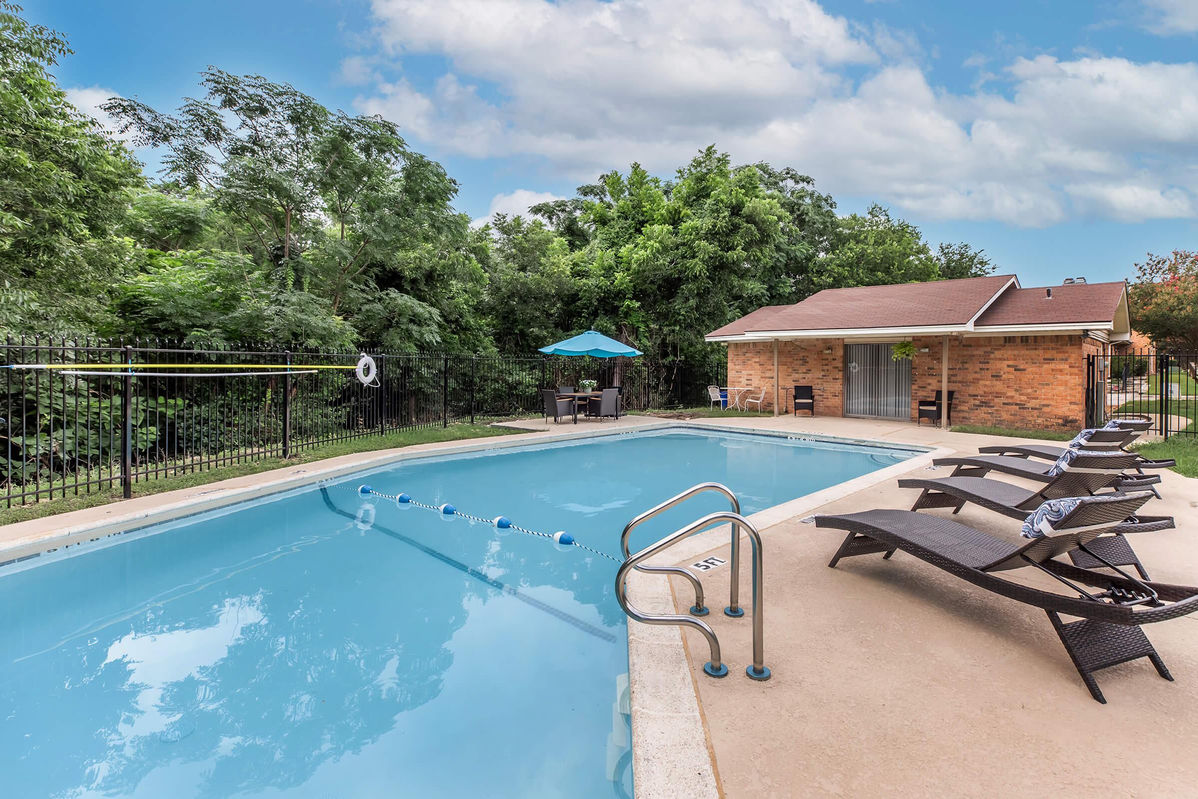 A well-maintained outdoor swimming pool surrounded by lounge chairs and a shaded seating area. Lush green trees line the background, and a small building is visible nearby. Bright blue skies with fluffy clouds add to the inviting atmosphere.
