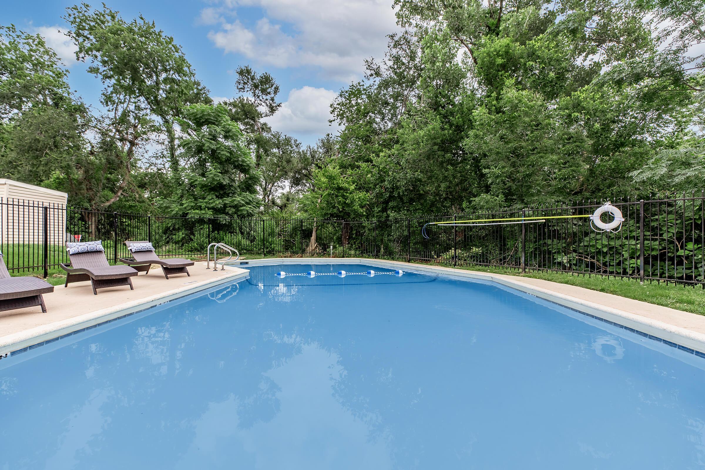A clear, blue swimming pool surrounded by a black metal fence, with several lounge chairs on the deck. Lush green trees and vegetation are visible in the background under a partly cloudy sky. A life ring is mounted on the fence near the pool.