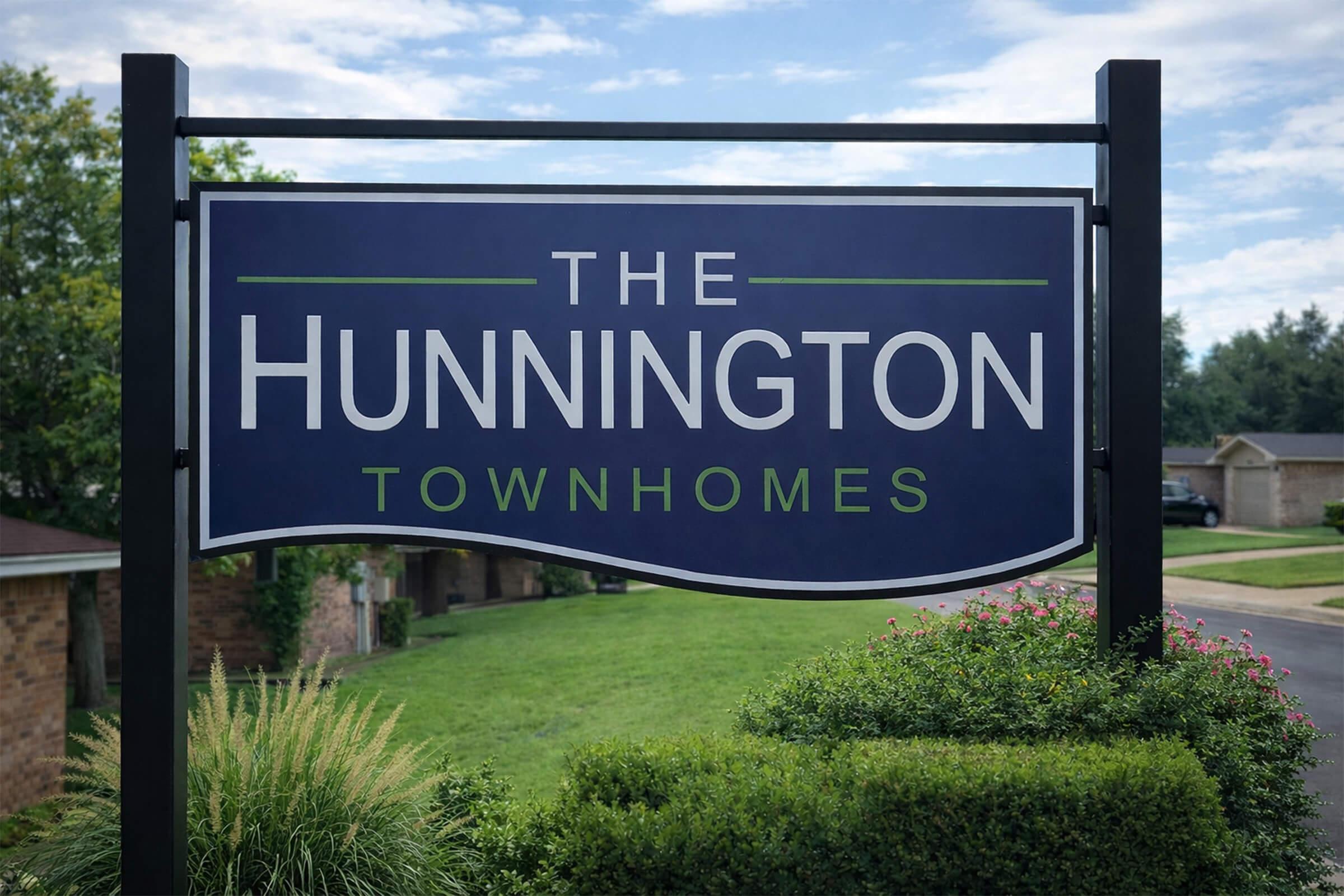 A sign for "The Huntington Townhomes" displayed prominently outside, featuring a blue background and white lettering with green accents. Surrounding greenery and a paved pathway are visible, suggesting a welcoming residential community. The sky is clear, indicating a pleasant day.