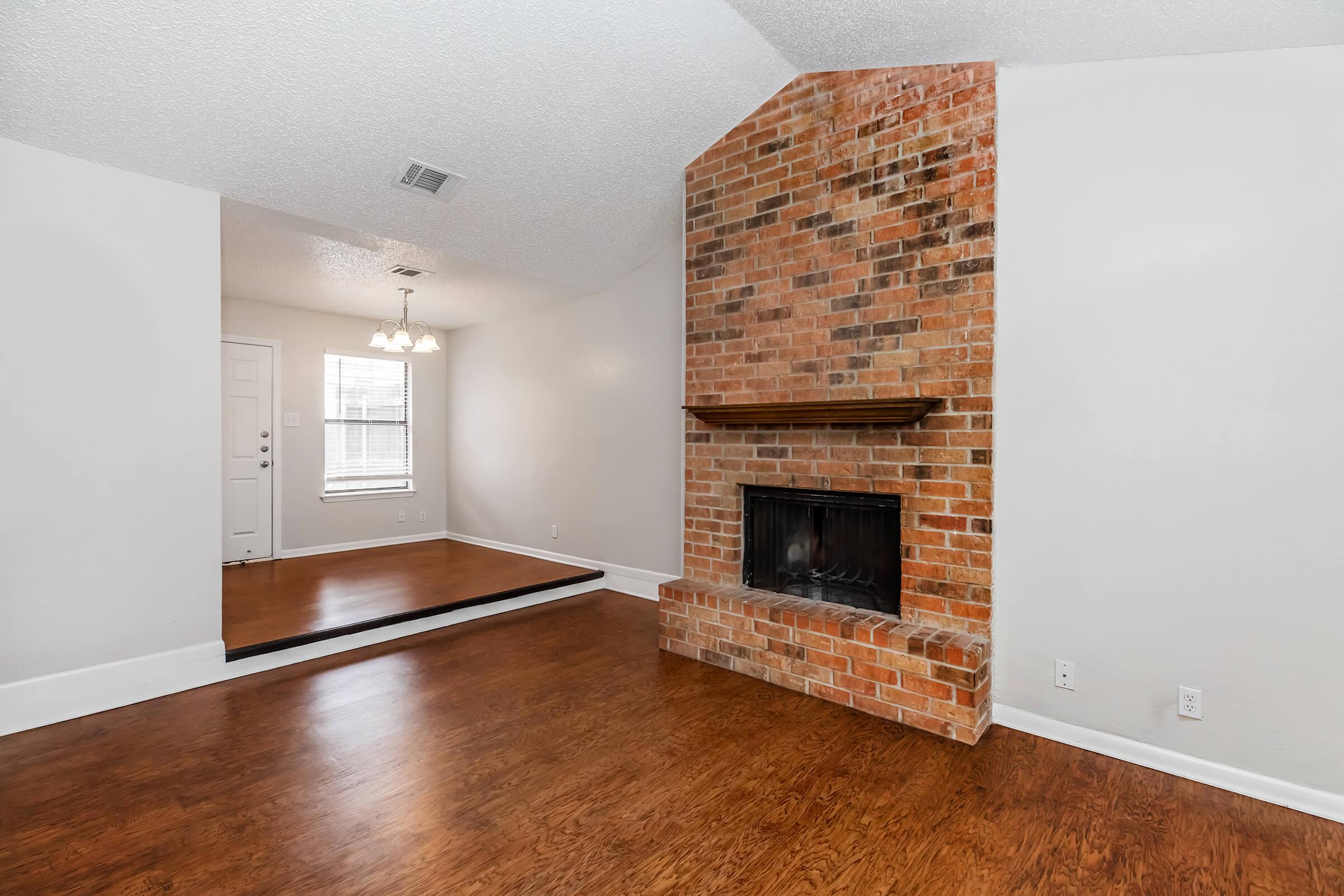 A spacious living room featuring a rustic brick fireplace as the focal point, hardwood floors, and light-colored walls. There is a doorway leading to another room, and a large window providing natural light. The overall ambiance is cozy and inviting.