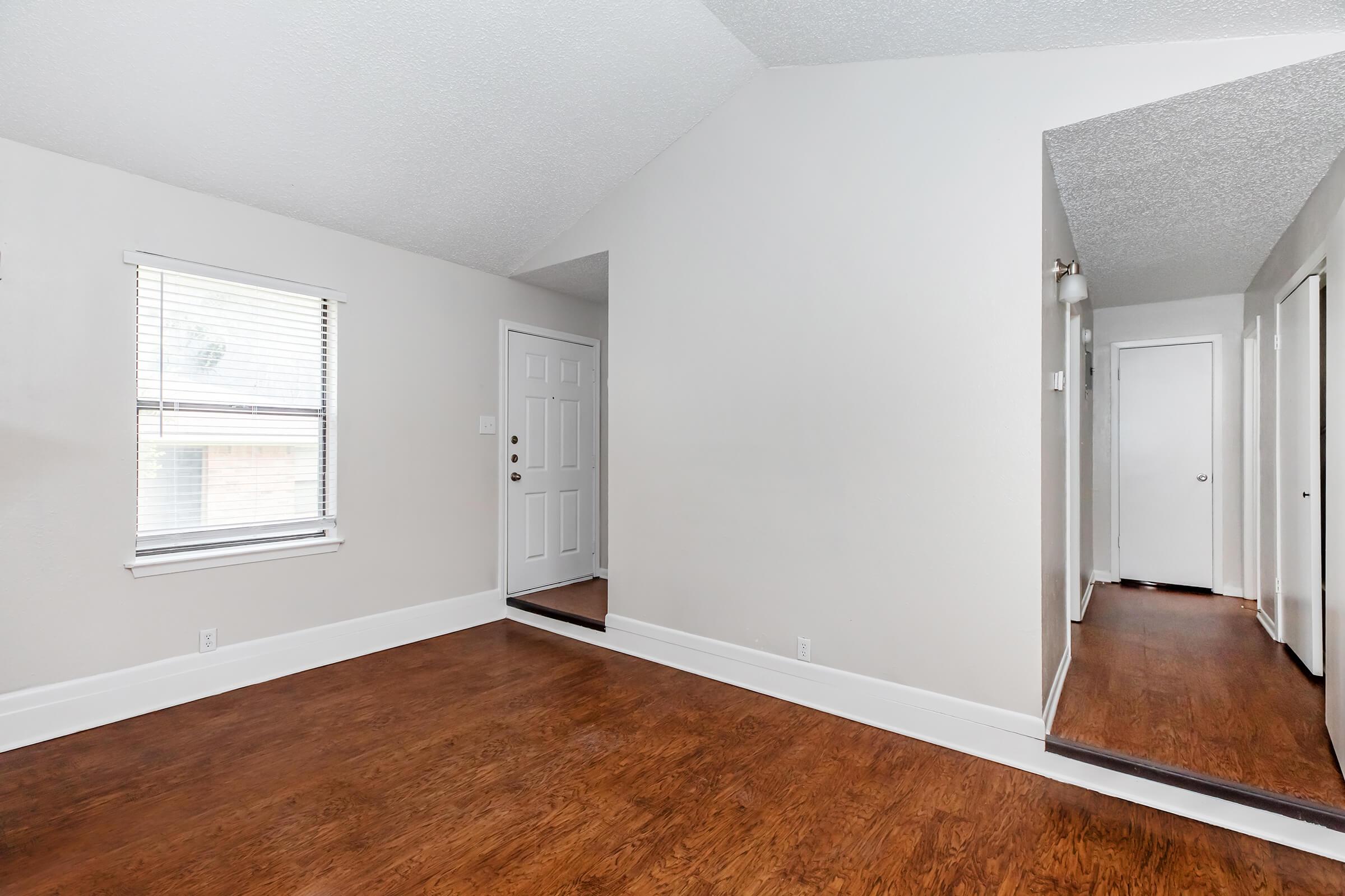 A well-lit living room with a high, angled ceiling and light-colored walls. There is a doorway leading to a hallway on the right, and a front door with a window to the left. The floor is made of dark wood, and the space appears clean and inviting, with natural light coming in from the window.