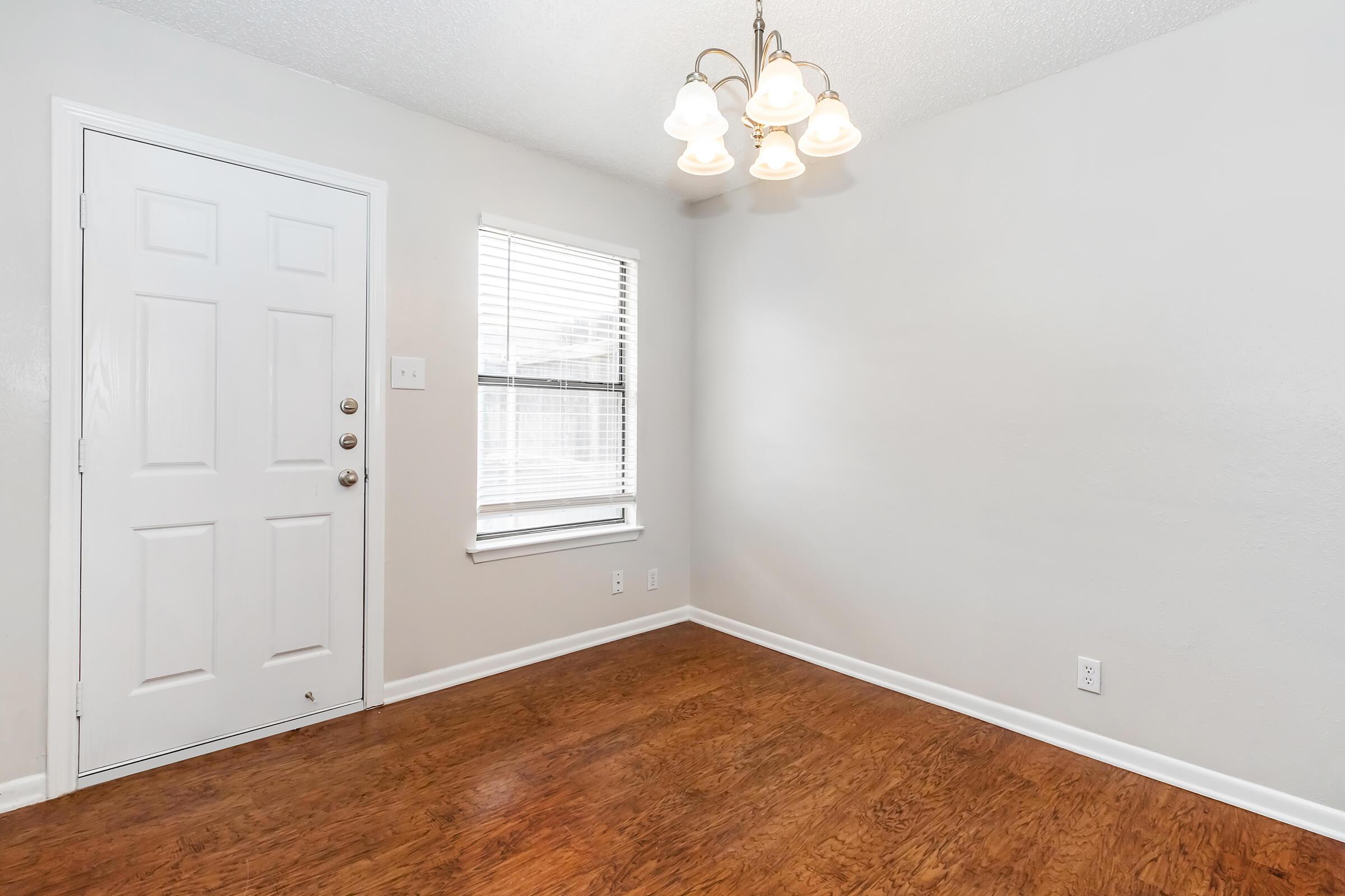 A small, empty room featuring a light fixture with multiple bulbs, a door on the left, and a window with blinds on the right. The walls are painted light gray, and the flooring is a warm, wooden texture. The space is well-lit and has a clean, minimalist appearance.