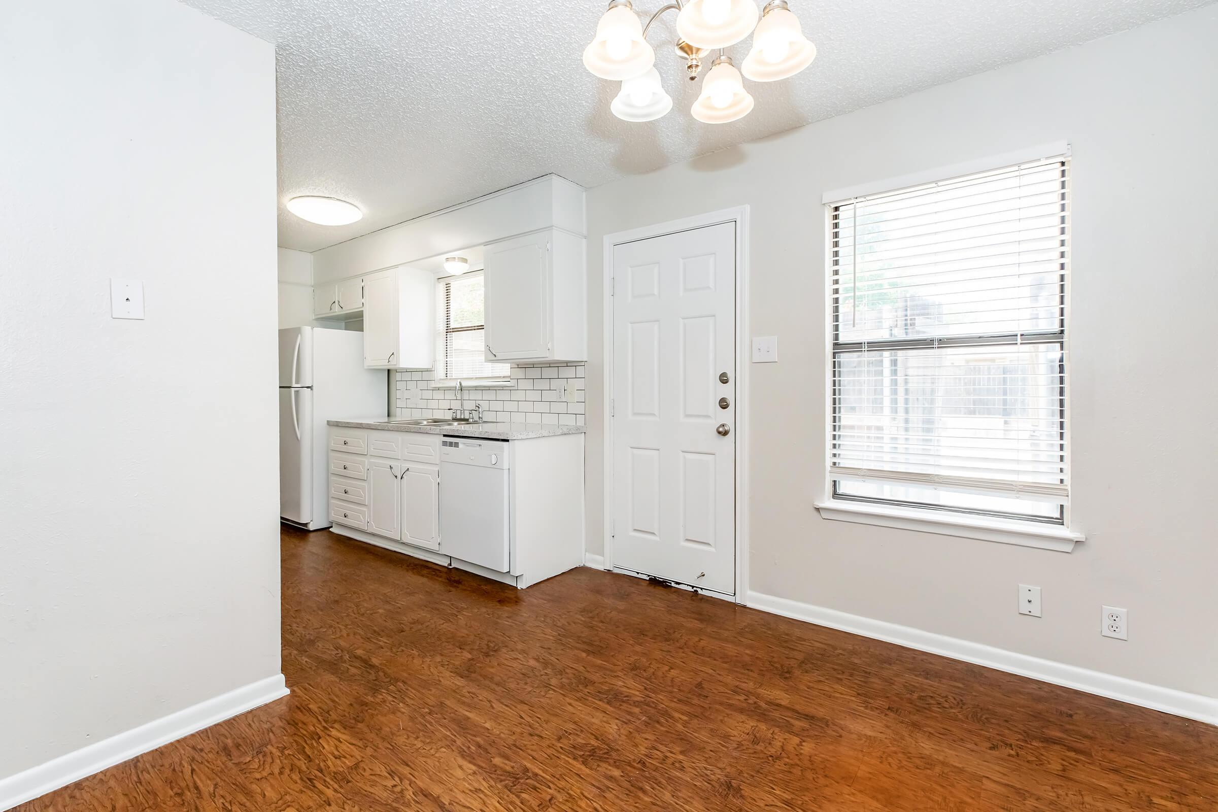 An interior view of a small apartment featuring a kitchen area on the left with white cabinetry and a sink. The living area has light-colored walls and a wooden floor. A door leads outside, and a window with blinds allows natural light into the space. The ceiling has a light fixture with multiple bulbs.