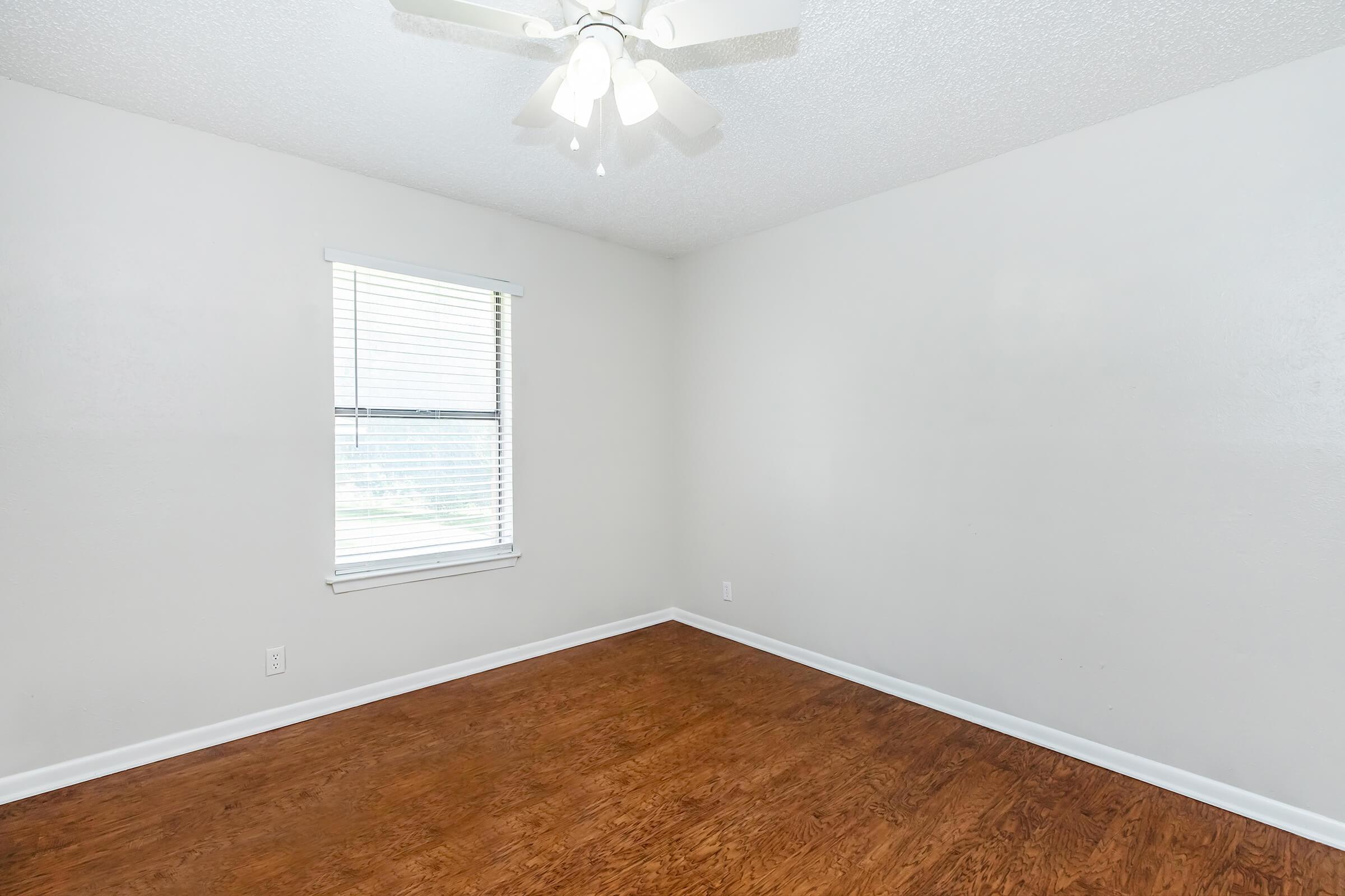 An empty room with light-colored walls and a wooden floor. A ceiling fan with lights is visible, and there is a single window with blinds letting in natural light. The room is devoid of furniture or decorations, creating a minimalist and spacious appearance.