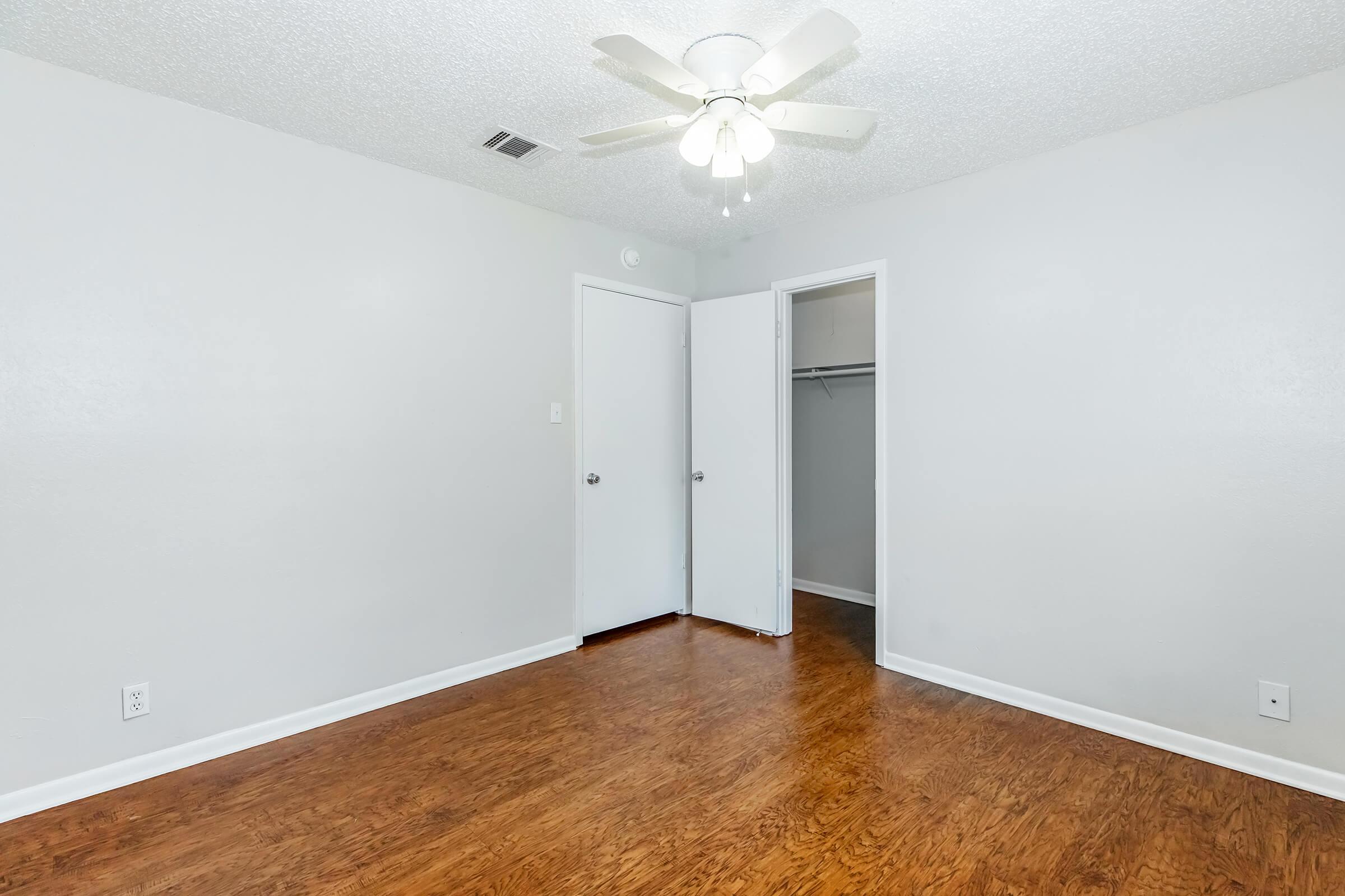A spacious, empty bedroom featuring light-colored walls and a ceiling fan. The floor is made of hardwood, and there's a closed closet visible with sliding doors. Two doors lead to different areas, creating a simple and clean aesthetic. Natural light brightens the room.