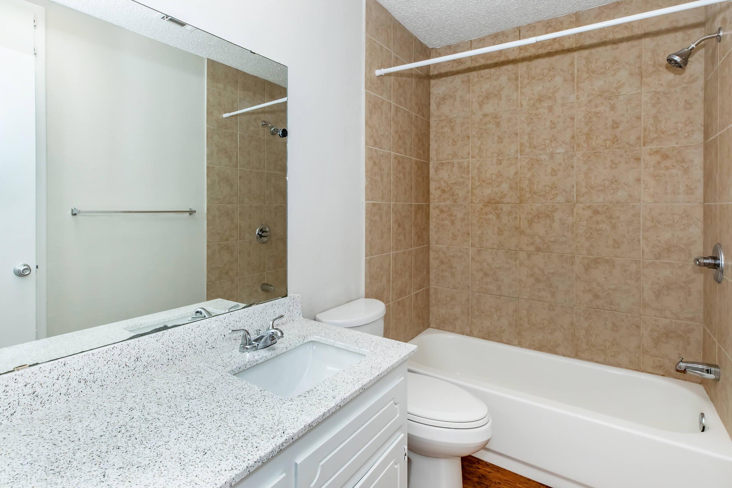 A clean and modern bathroom featuring a bathtub with a shower, a wall-mounted mirror above a vanity with a sink, and beige tiled walls. The space has bright lighting and a minimalistic design, creating a fresh and inviting atmosphere.