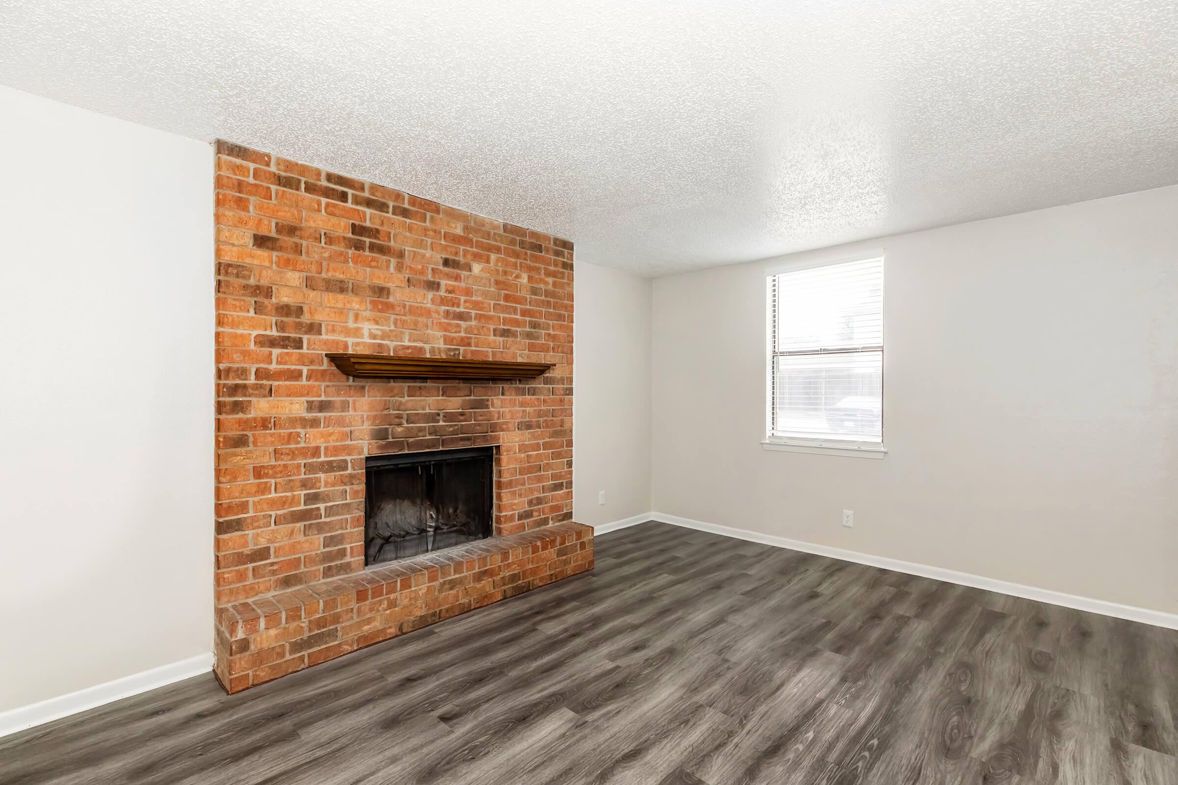 Living room featuring a simple design with a brick fireplace, a wooden mantel, and a light-colored wall. The flooring is dark wood laminate, and a window provides natural light, enhancing the spacious feel of the room.