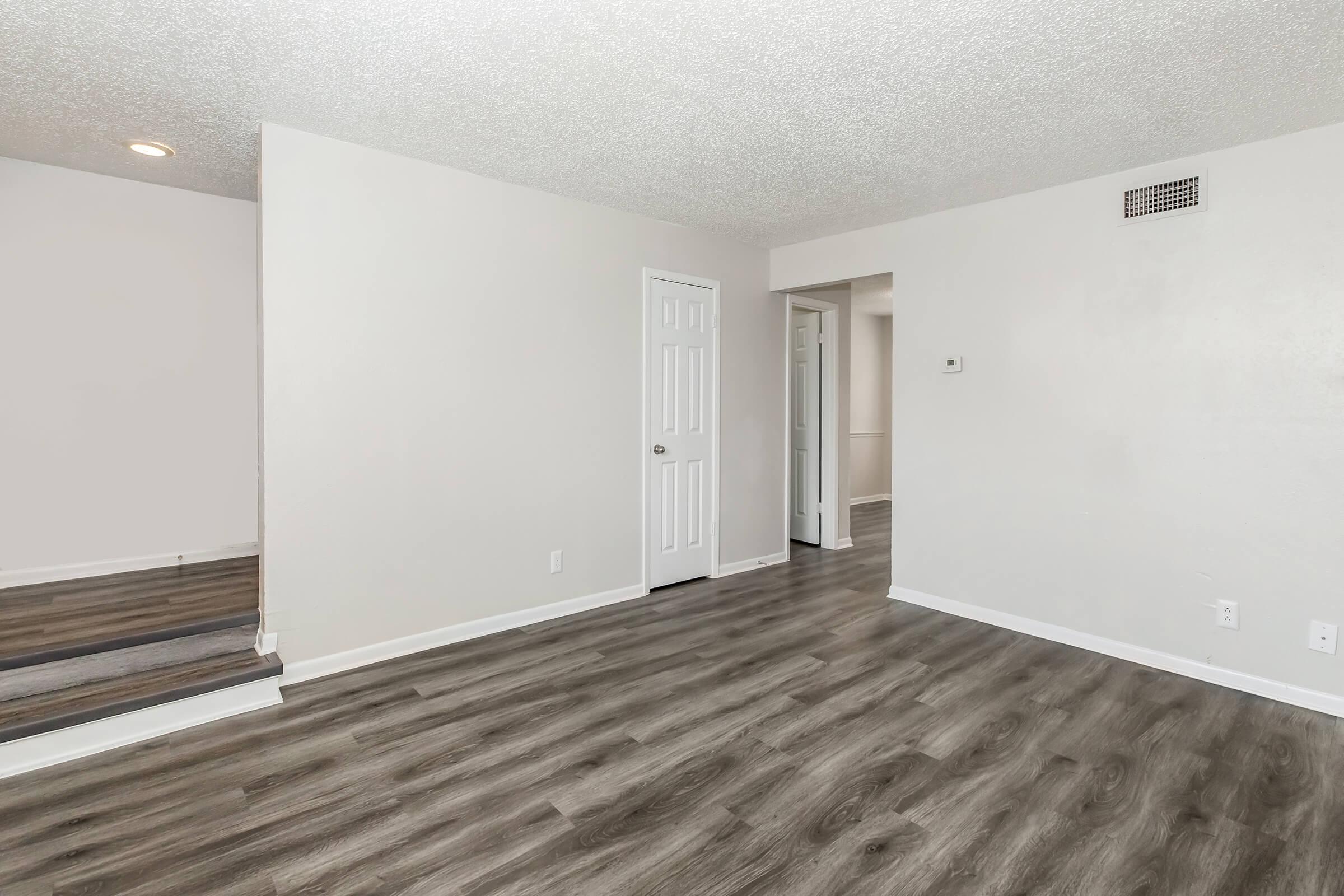 A spacious, brightly lit living area with light-colored walls and modern laminate flooring. On the left, there is a doorway leading to another room, and a hallway visible in the background. The overall atmosphere is clean and inviting, suitable for various decorating styles.