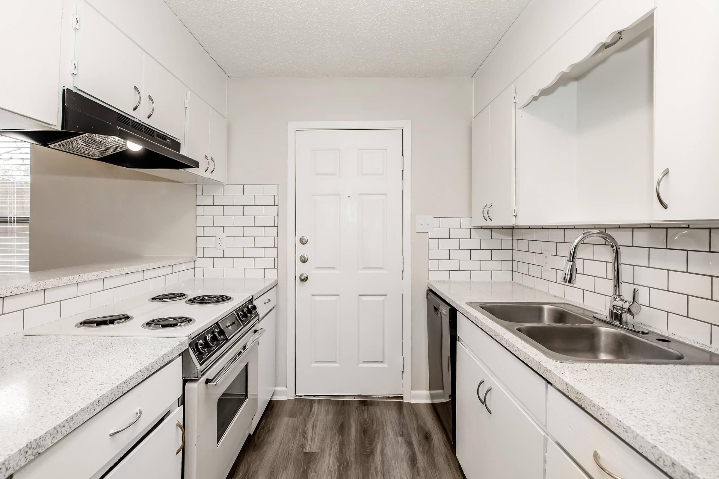 Modern kitchen featuring a gas stove, oven, and dual sinks with a stainless steel faucet. The walls are adorned with white subway tiles, and the cabinetry is sleek and white. A door leads outside, and the flooring is a warm wood-style finish, creating an inviting atmosphere.