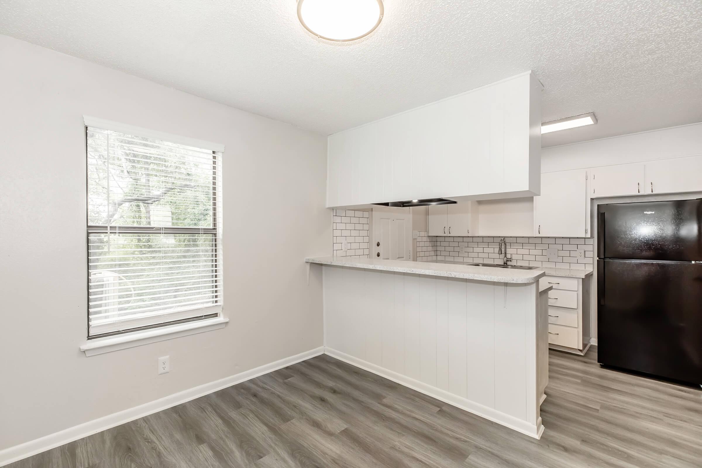 A modern kitchen featuring white cabinetry, a granite countertop, and a black refrigerator. There are large windows allowing natural light, and the flooring is a light gray vinyl. The layout includes a bar area overlooking the living space.
