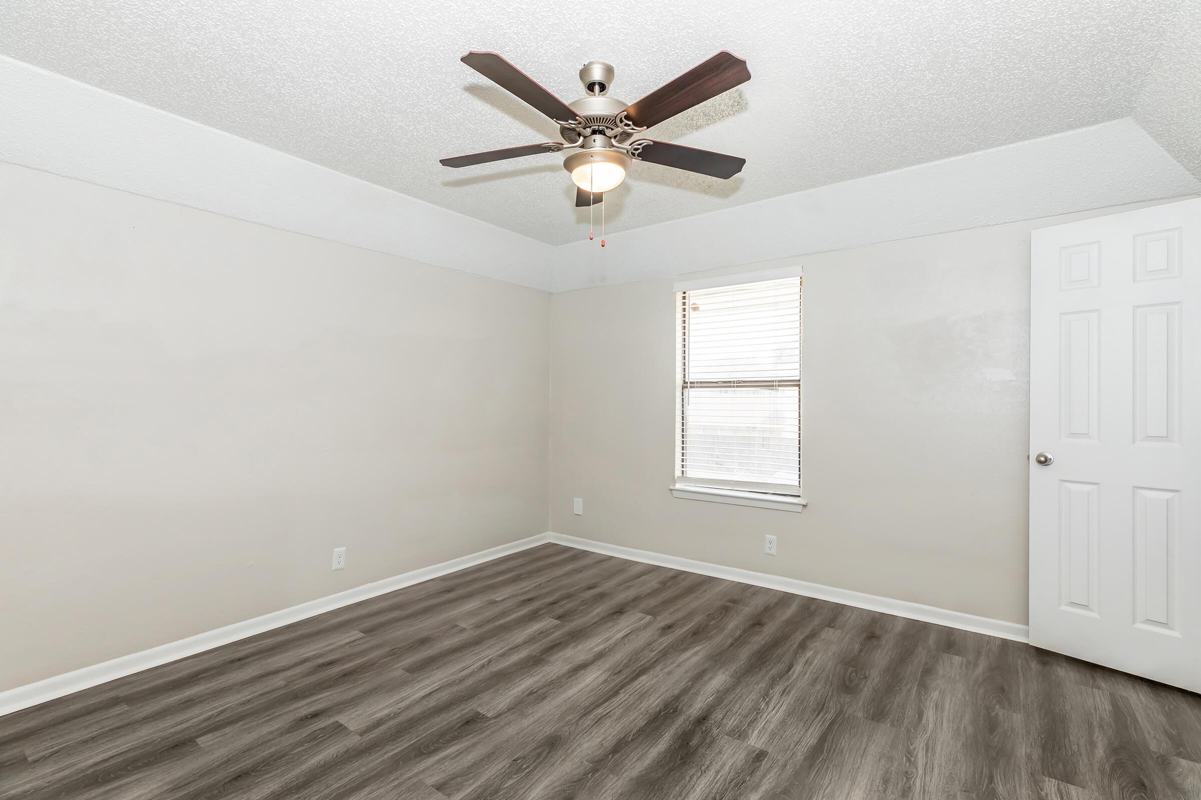 Empty room with light-colored walls and a ceiling fan. The floor features laminate flooring, and there is a window with white blinds. A white door is visible on the right side of the image. The room has a clean and minimalistic appearance, with ample natural light.