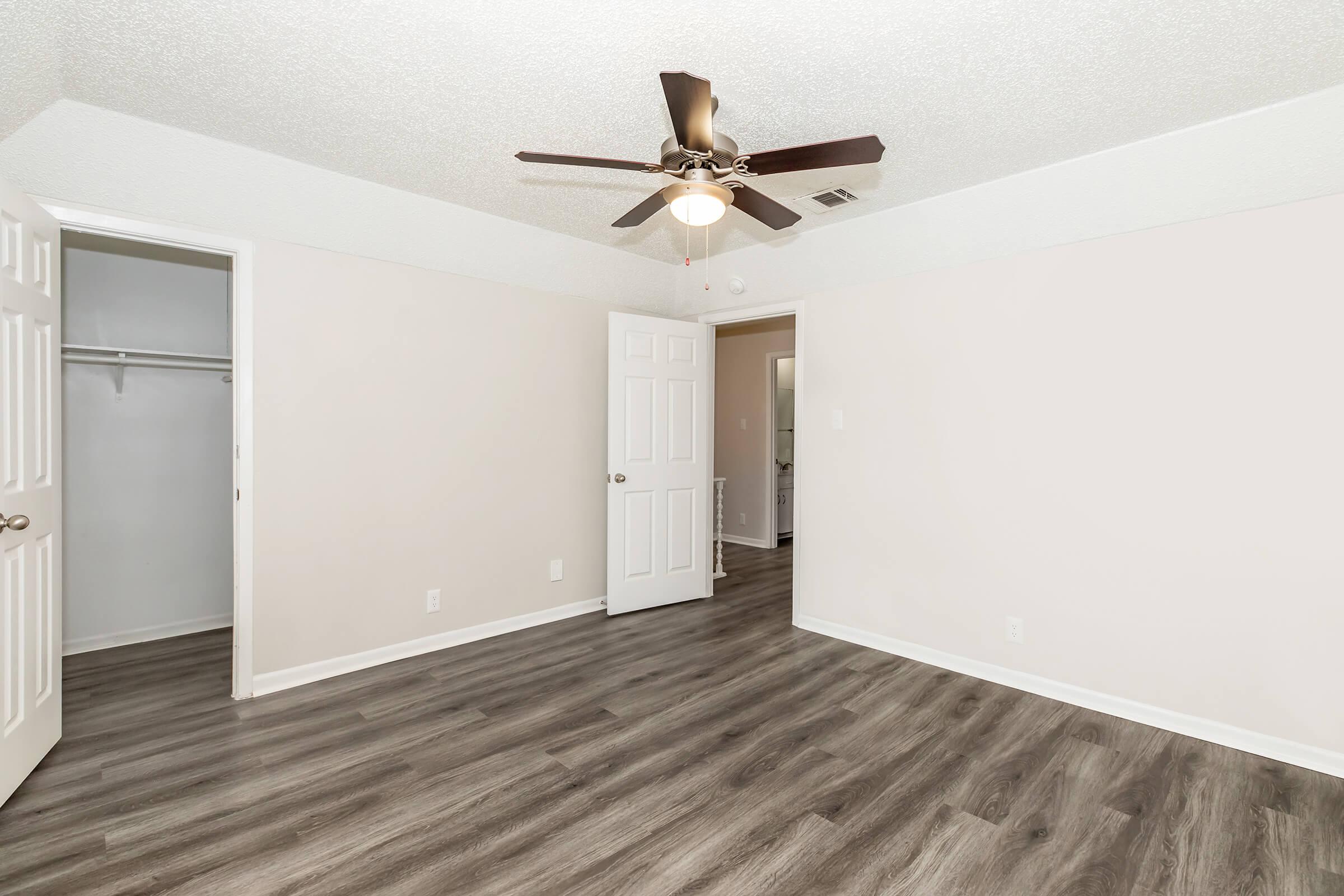 Empty room with light-colored walls, a ceiling fan, and dark wood-style flooring. One door leads to a small closet, while another door is open to a bathroom. The space is well-lit and unfurnished, creating a blank canvas for potential furniture and decor.