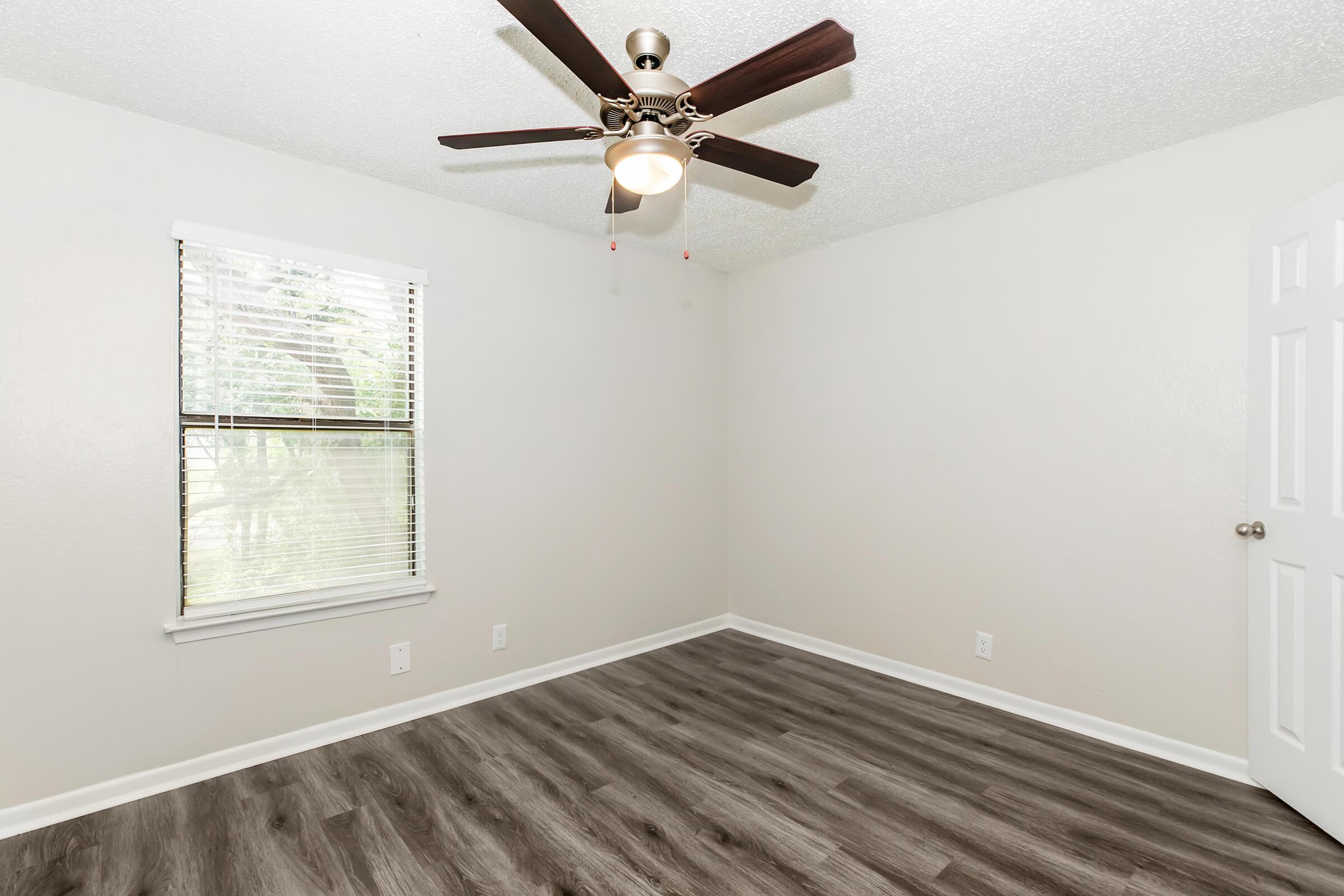 Empty room with light-colored walls and a ceiling fan with wooden blades. The flooring is dark wood laminate. A window with blinds allows natural light, and there's a closed white door on the right side of the image.