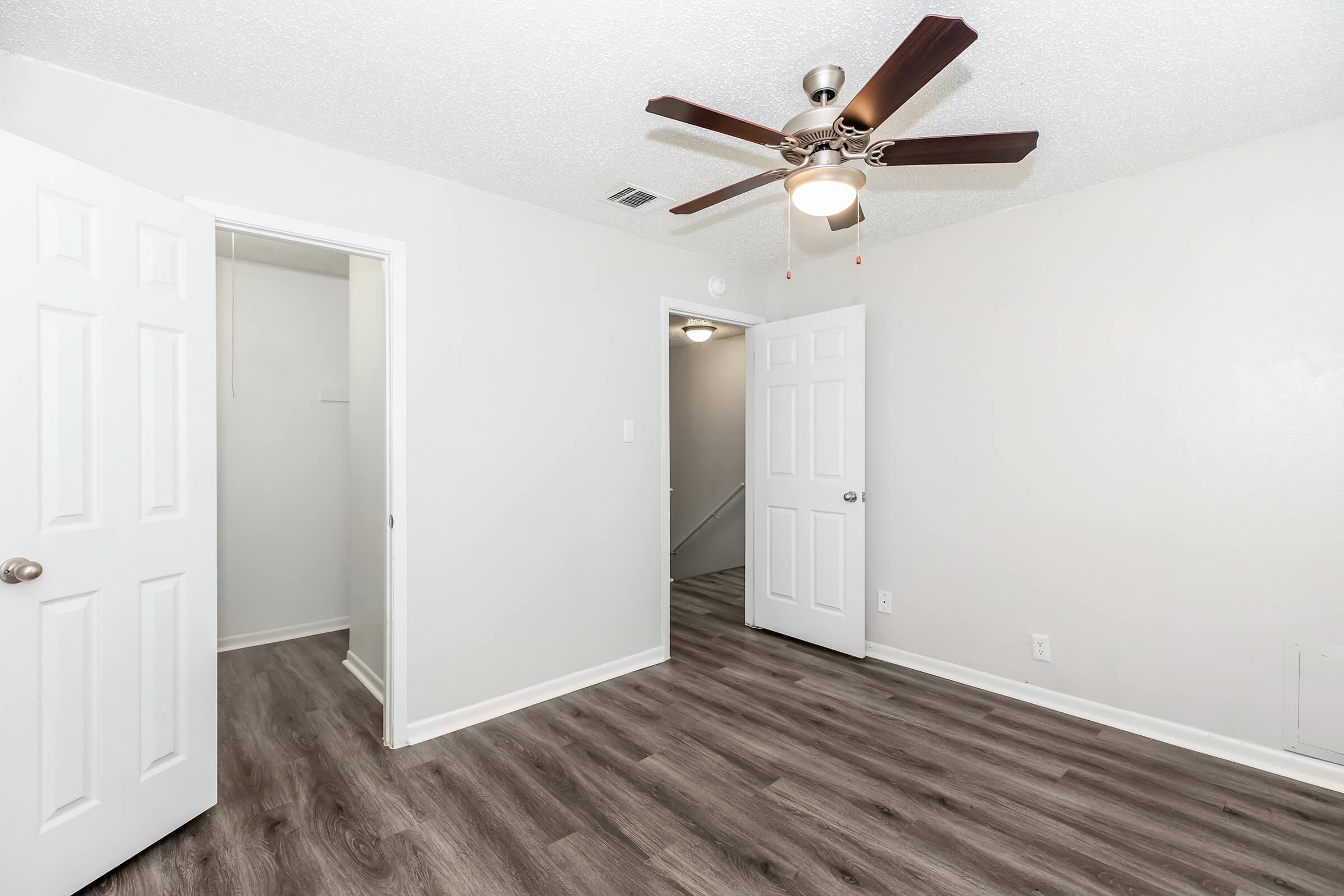 A well-lit room featuring laminate flooring, a ceiling fan, and two open white doors leading to adjacent spaces. The walls are painted light gray, creating a clean and modern atmosphere. The room is empty, suggesting it could be used as a bedroom or office.