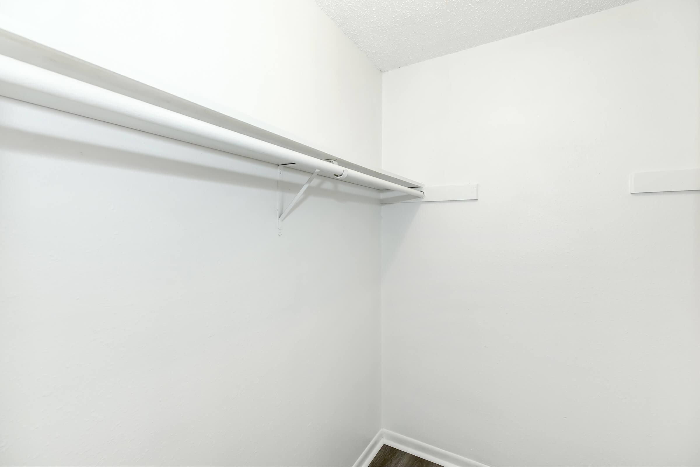 Empty white closet with a single hanging rod against the wall, featuring a plain white interior and wooden flooring. The space is well-lit and minimalistic, with no items or shelves present.