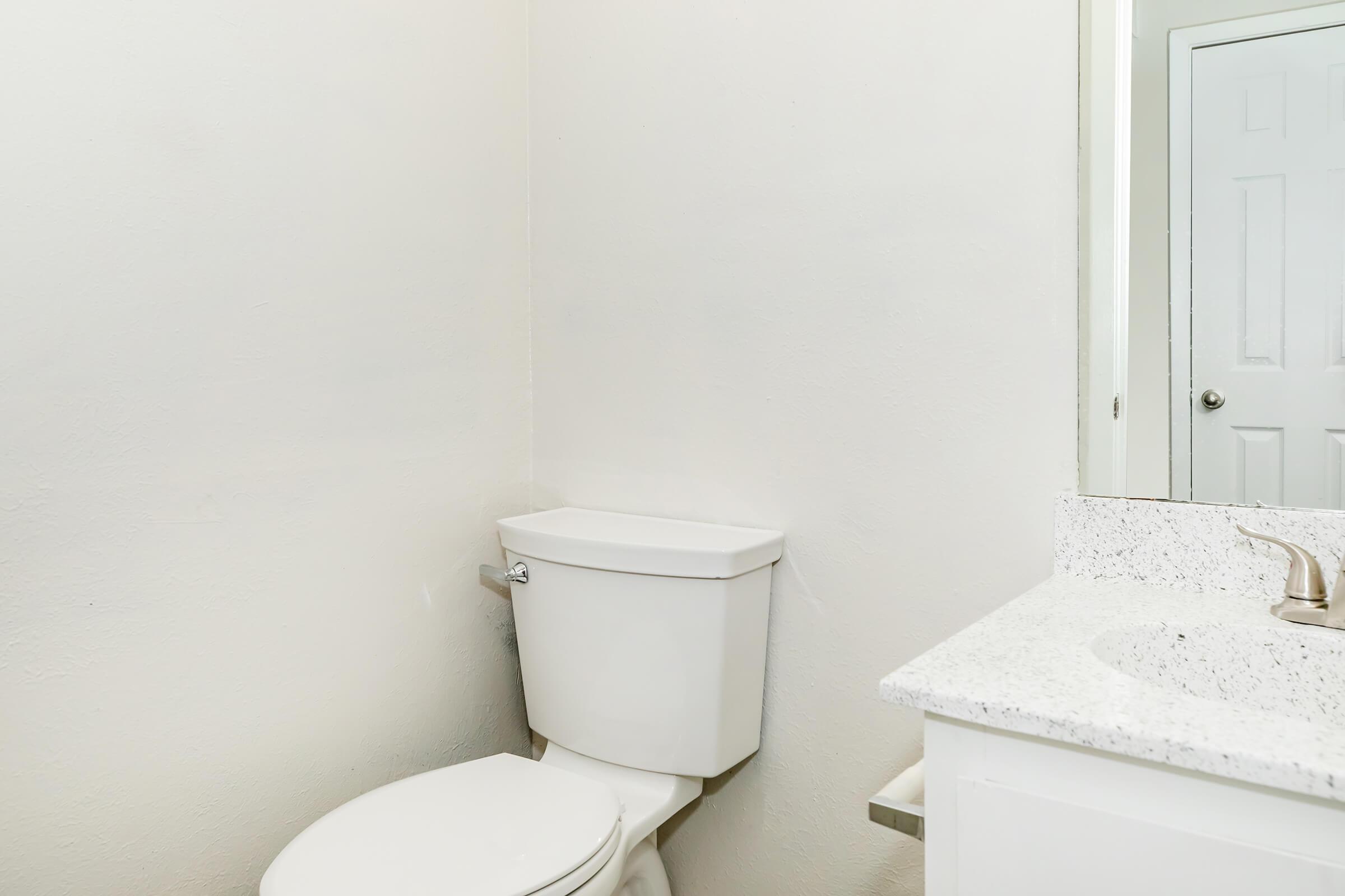 A clean, minimalist bathroom featuring a white toilet and a countertop with a sink. The walls are plain and light-colored, and a mirror is reflected in the background. The overall space is bright and uncluttered, emphasizing simplicity.