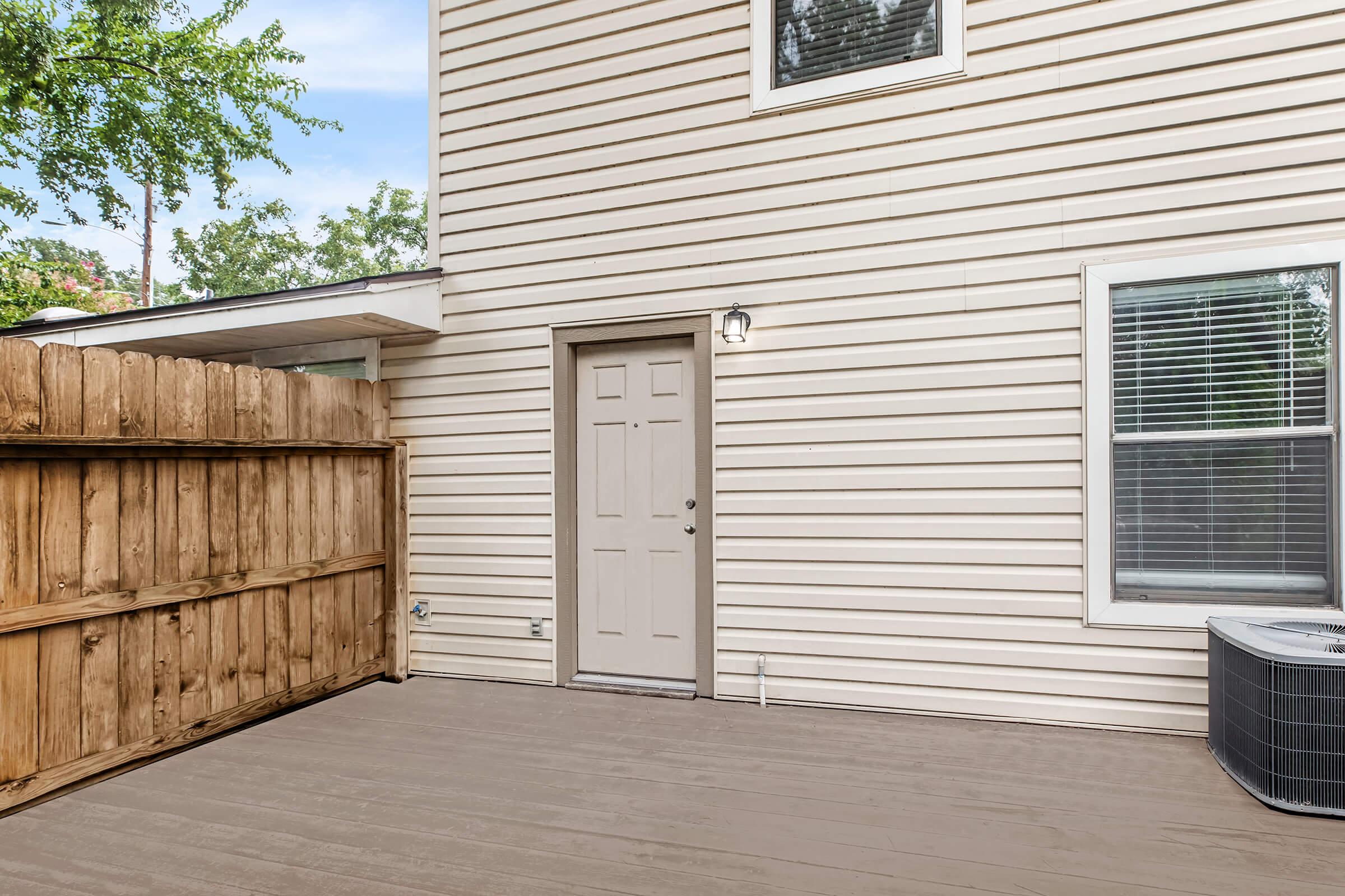 A view of the exterior of a house featuring a wooden fence, a door with a light fixture beside it, and a window. The wall has a light beige siding, and there's an air conditioning unit on the ground. Trees and greenery can be seen in the background.
