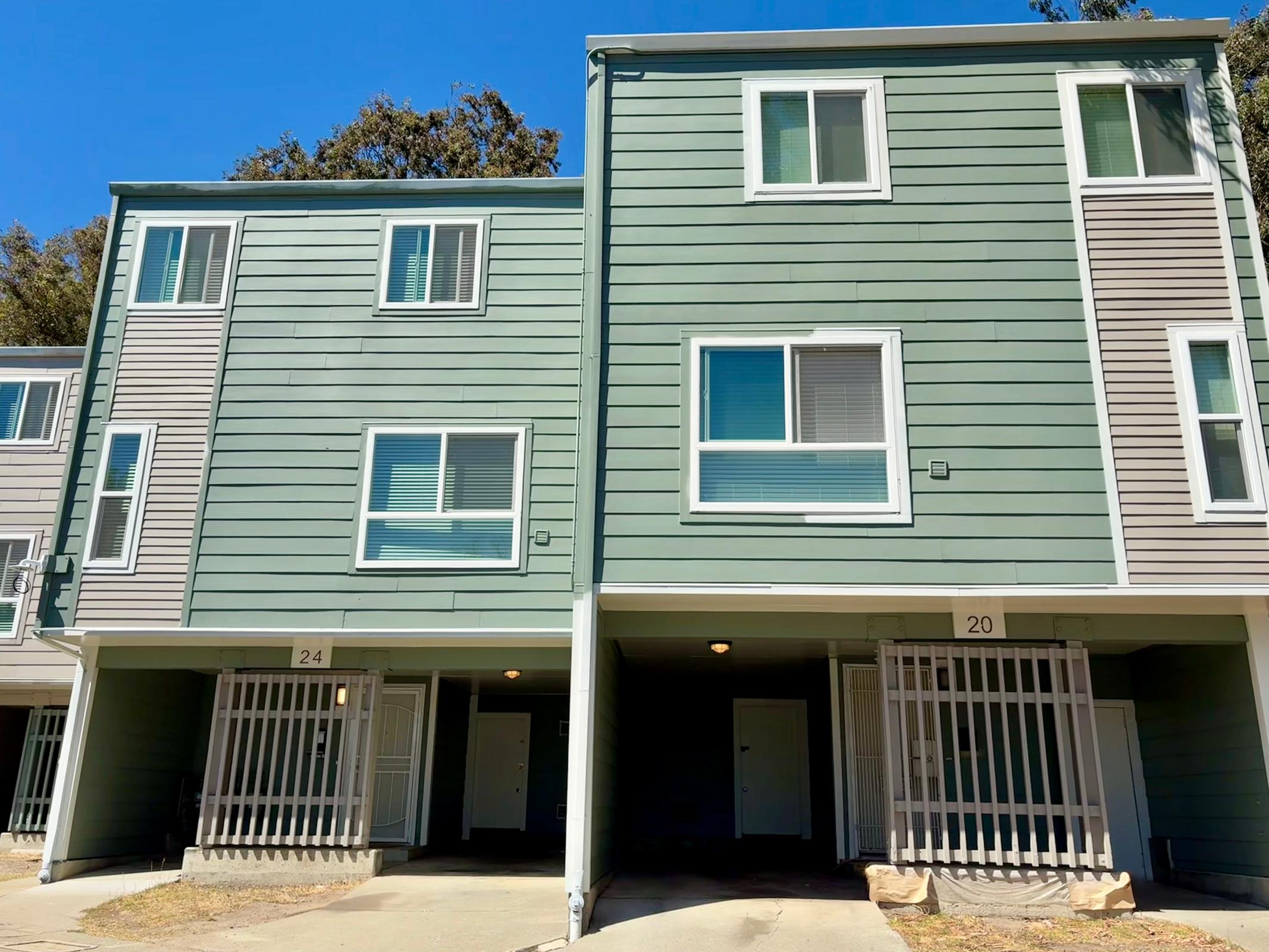 Two modern, multi-story apartment buildings with green siding. The buildings have multiple windows and doors, with unit numbers 24 and 20 visible. Each entrance is protected by a metal gate, and the ground is paved. The bright blue sky provides a clear backdrop.