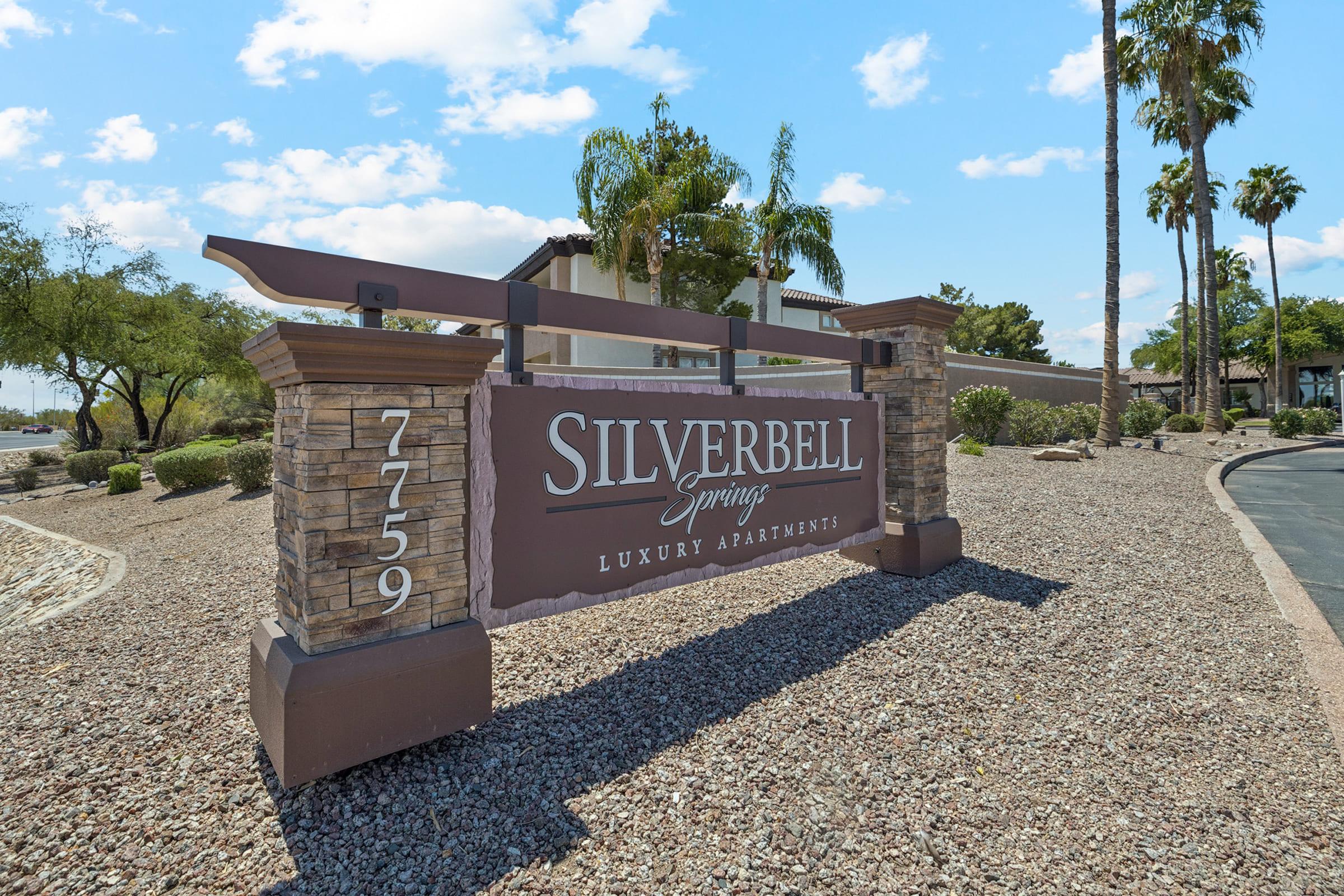 Sign for Silverbell Springs Luxury Apartments at 7759 with a decorative stone base, surrounded by landscaping and palm trees under a blue sky with scattered clouds.