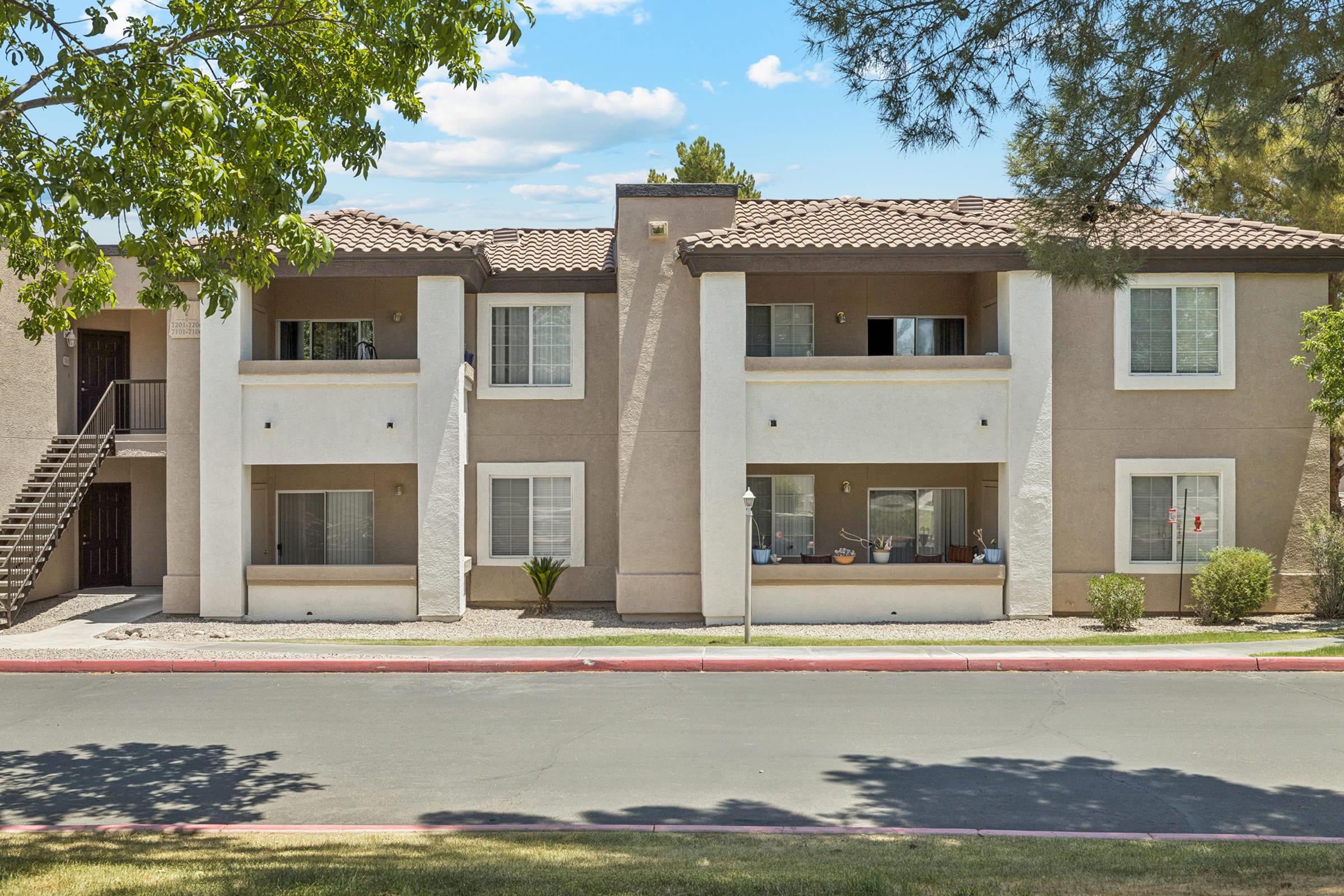 A modern two-story apartment building with a light-colored exterior and a tiled roof. There are multiple units visible, each with windows and balconies. The landscape includes a small lawn and a tree in the foreground, and a paved road runs in front of the building under a clear blue sky.