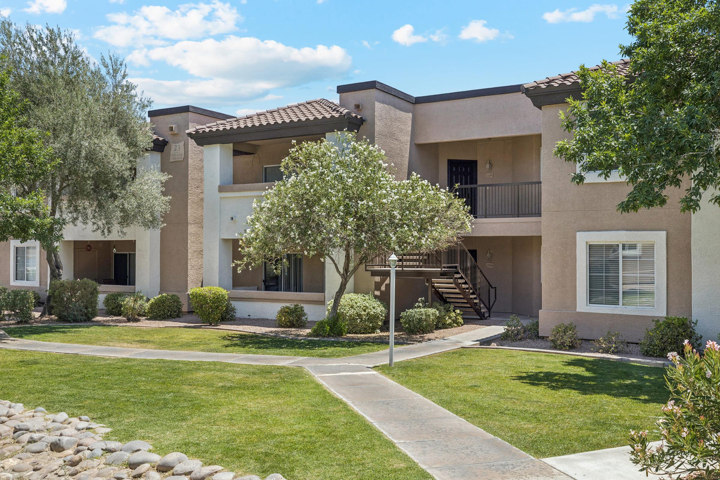 A landscaped courtyard featuring a multi-unit apartment building. The building has two levels with balconies, surrounded by greenery, including trees and shrubs. A pathway leads through the lawn, providing access to the building and highlighting a sunny blue sky with scattered clouds.