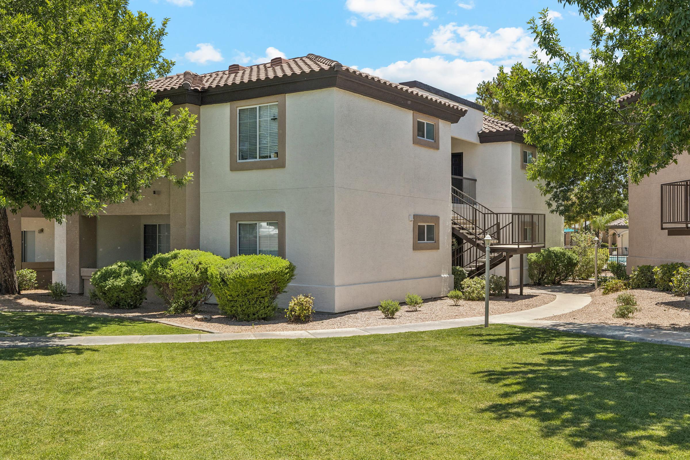 A two-story residential building surrounded by well-maintained green lawns and shrubs. The exterior features light-colored walls and a tiled roof. There are trees providing shade and a pathway leading to the entrance. The scene is bright and sunny, depicting a pleasant outdoor environment.