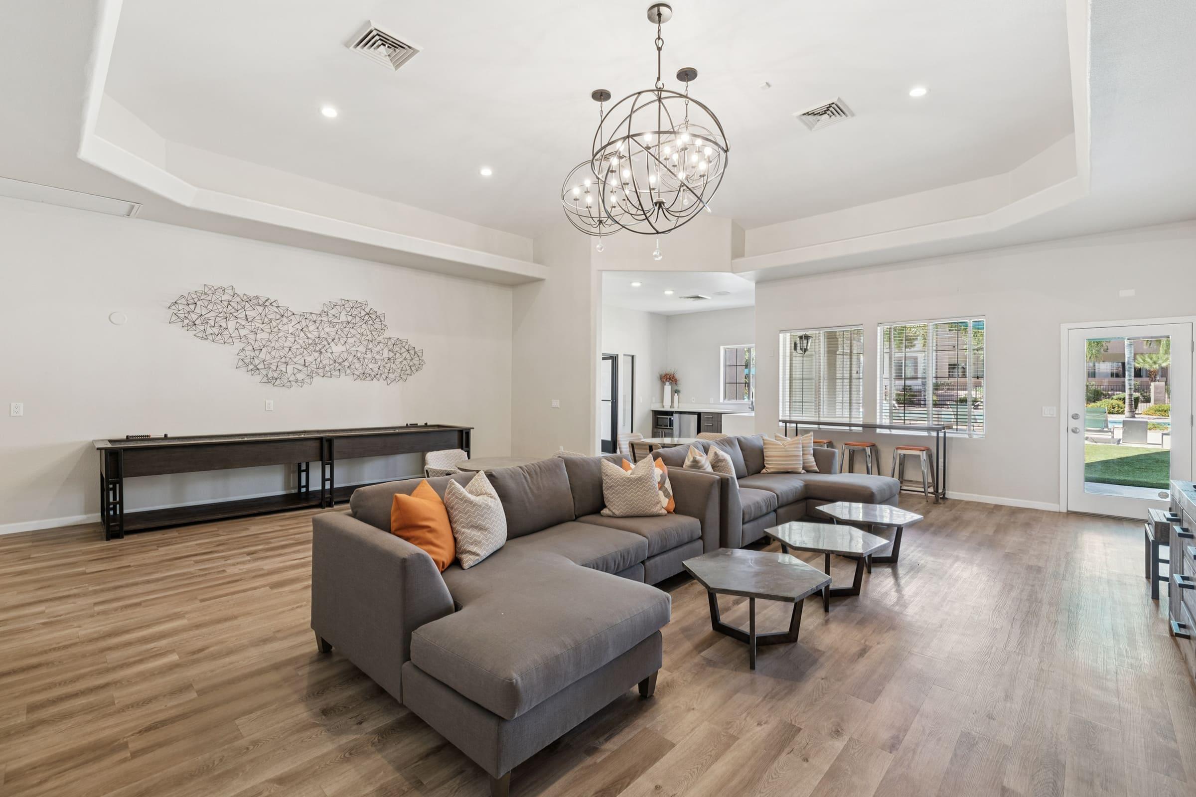 A modern living area featuring a spacious sectional sofa with decorative pillows, coffee tables, and large windows. A stylish chandelier hangs from the ceiling, while a wall art piece adds a contemporary touch. The room has warm wood flooring and is brightly lit, creating an inviting atmosphere.
