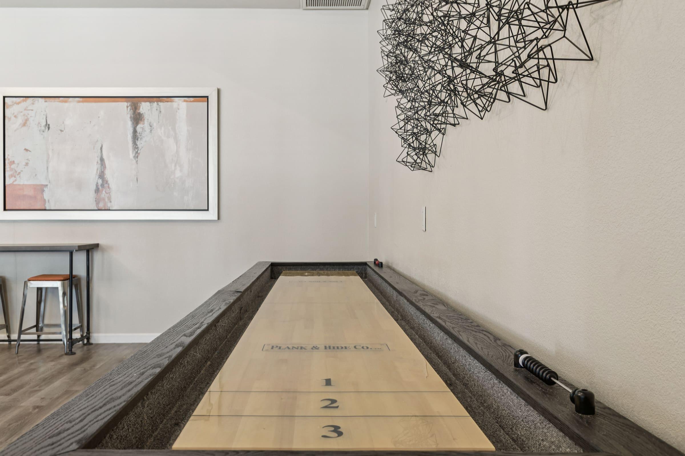 A wooden shuffleboard table is positioned in a modern interior with a light-colored wall. Above the table, there is a contemporary wire sculpture, and a framed abstract artwork hangs nearby. A small bar table with stools is visible in the background, adding to the stylish ambiance of the space.