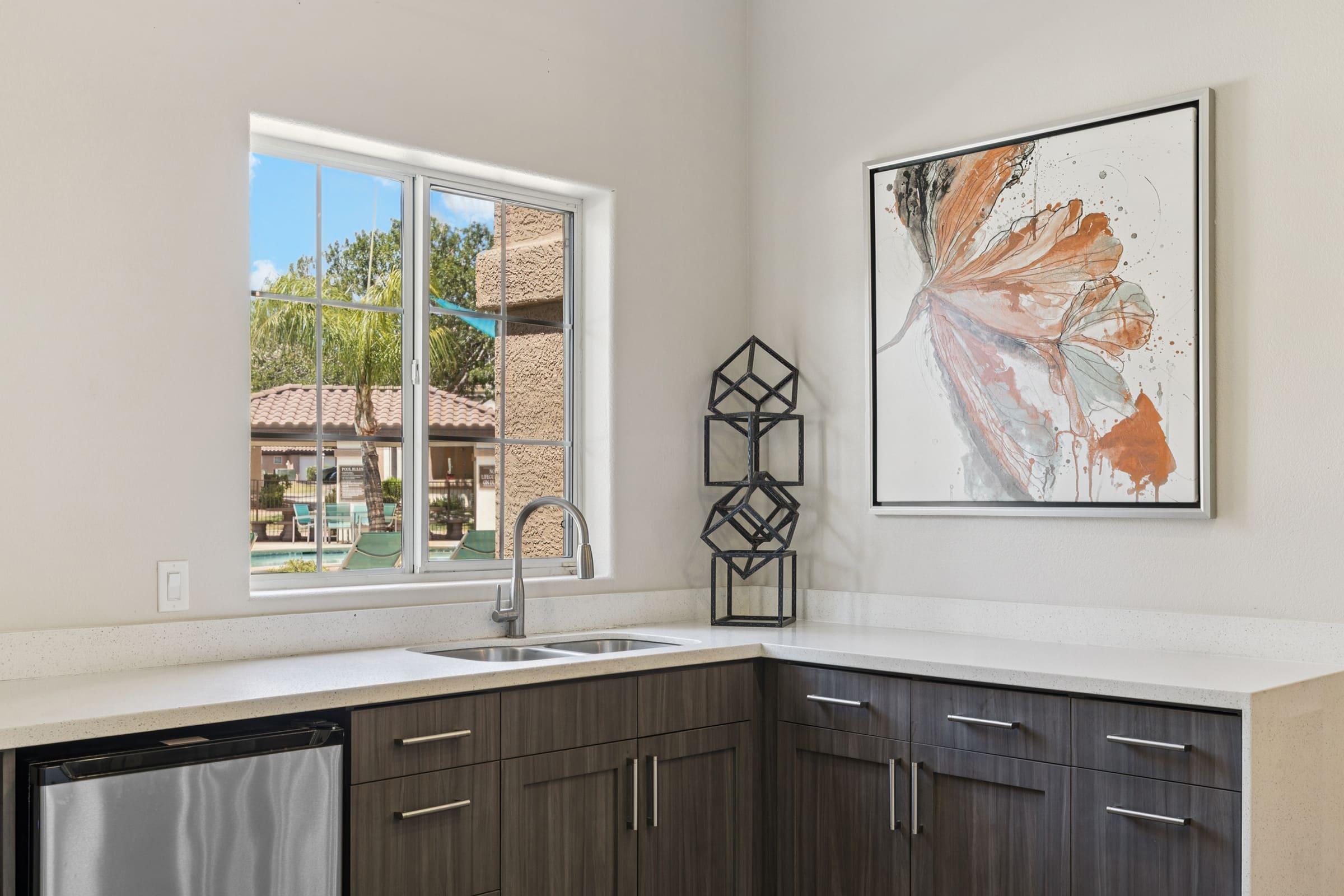 Modern kitchen with a sleek dark wood cabinetry, stainless steel sink and appliances. A large window offers a view of outdoor greenery, while a framed artwork featuring abstract floral designs hangs on the wall. A geometric sculpture adds a stylish touch to the countertop.
