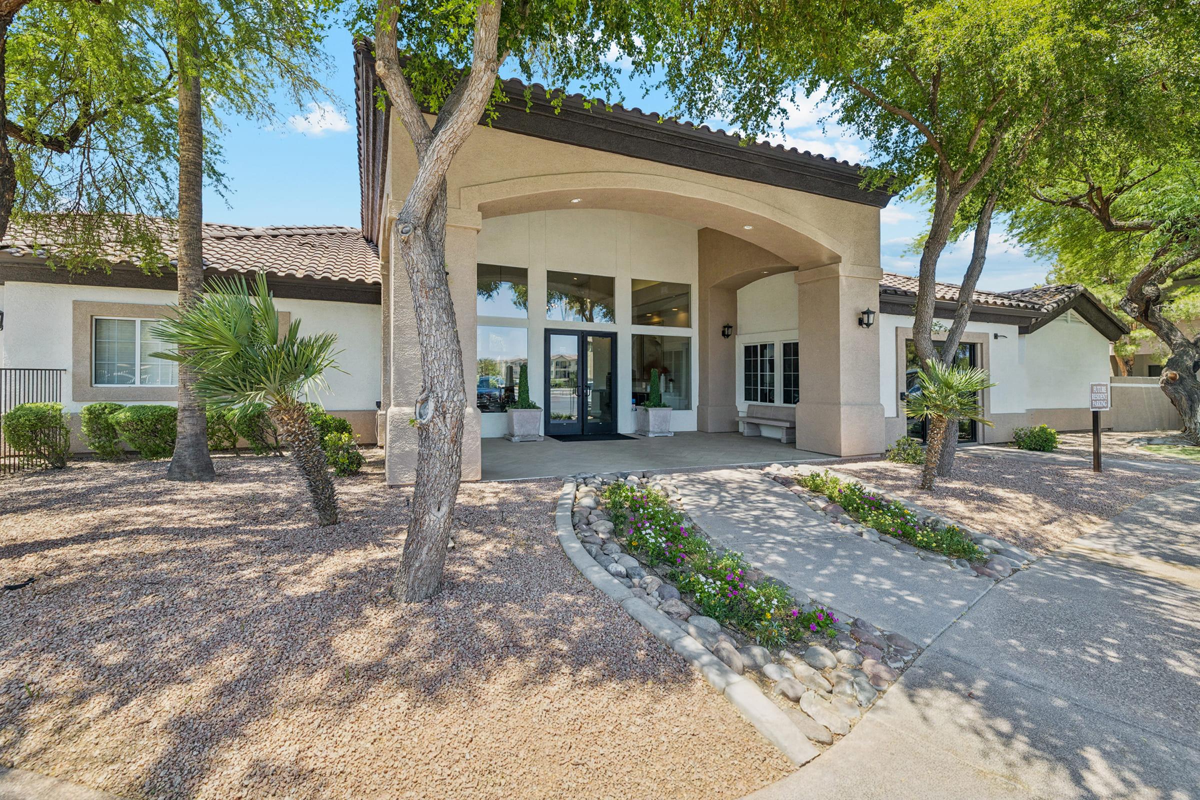 A modern building entrance surrounded by landscaped gardens, featuring a path lined with stones and flowers. Tall trees provide shade, and large windows enhance the welcoming appearance. The blue sky and bright sunlight create a cheerful atmosphere.
