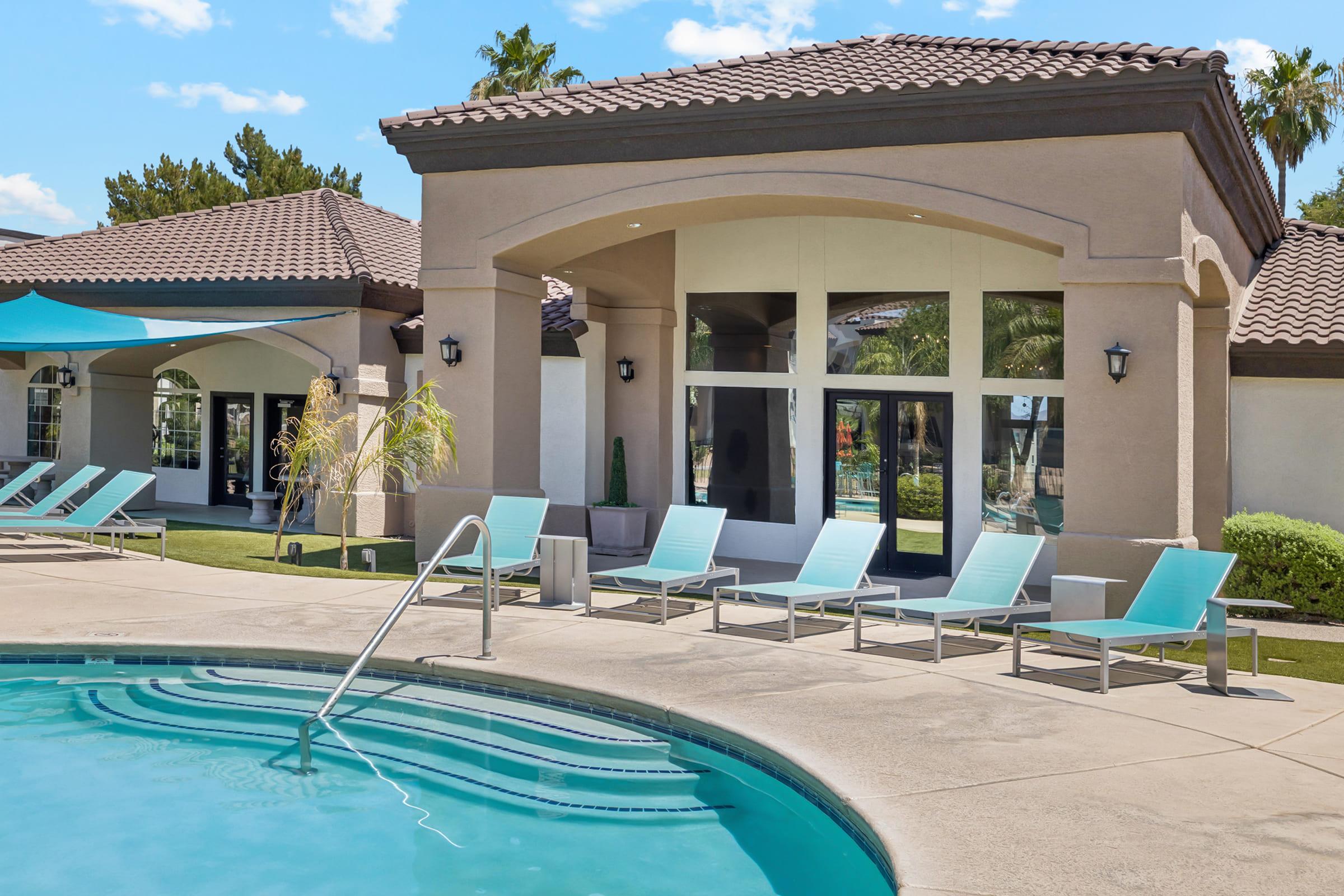 A modern outdoor pool area featuring a curved swimming pool with a gentle blue surface. Comfortable teal lounge chairs line the poolside, and a well-maintained building with large windows and a tiled roof is in the background, surrounded by palm trees and a sunny sky.