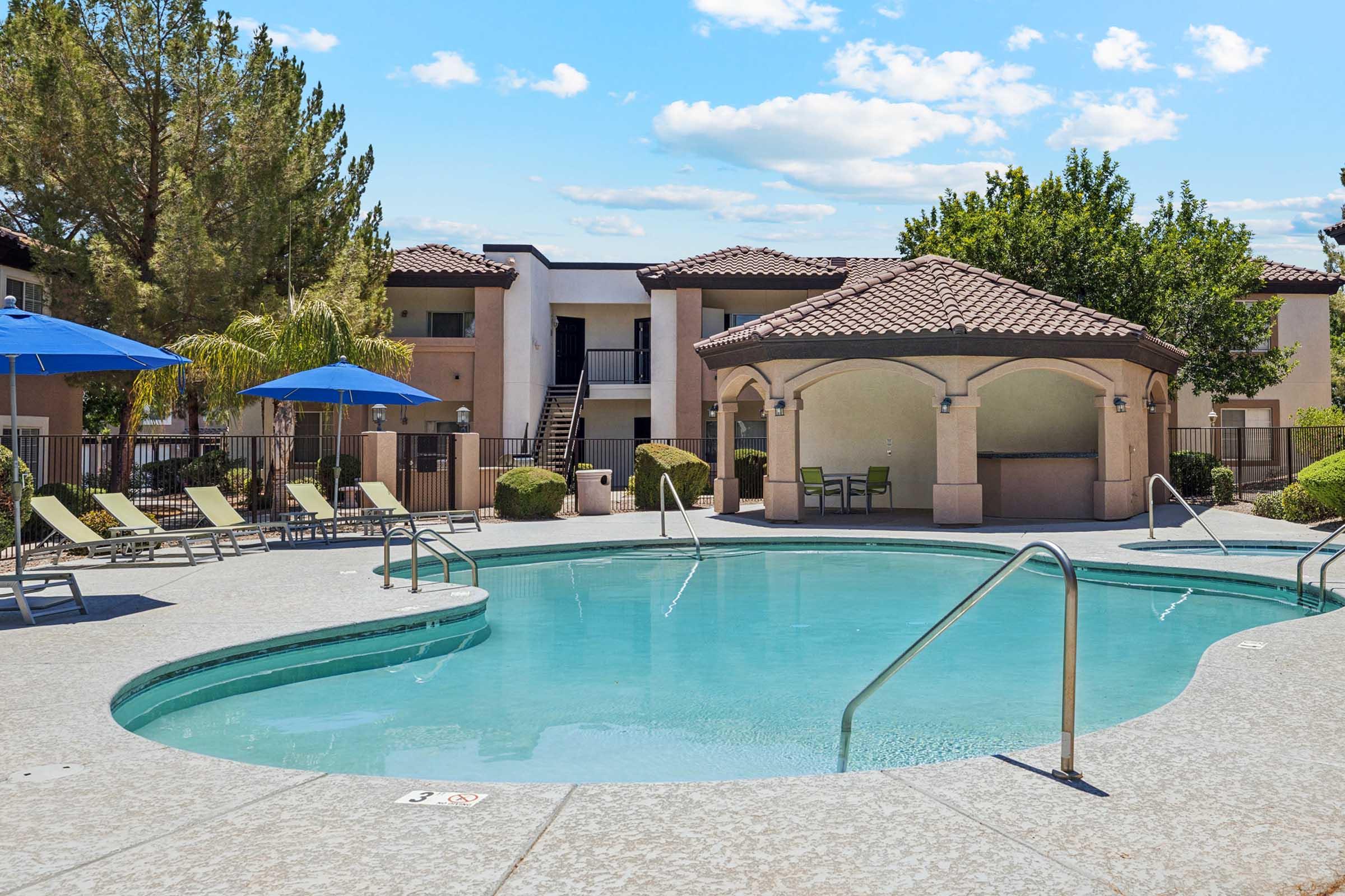 A tranquil swimming pool surrounded by lounge chairs and blue umbrellas, with a shaded gazebo nearby. Apartment buildings in the background are framed by green trees and a bright blue sky with fluffy clouds.