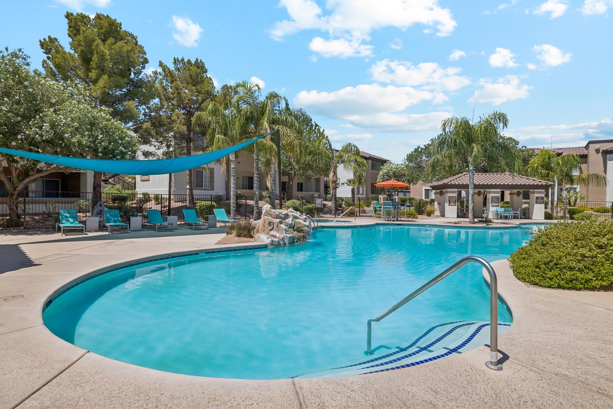 A serene swimming pool surrounded by lush greenery and palm trees. Lounge chairs are set up around the pool area, and a shaded sitting area with an umbrella can be seen. The sky is bright with fluffy clouds, creating a relaxing atmosphere for leisure and enjoyment.
