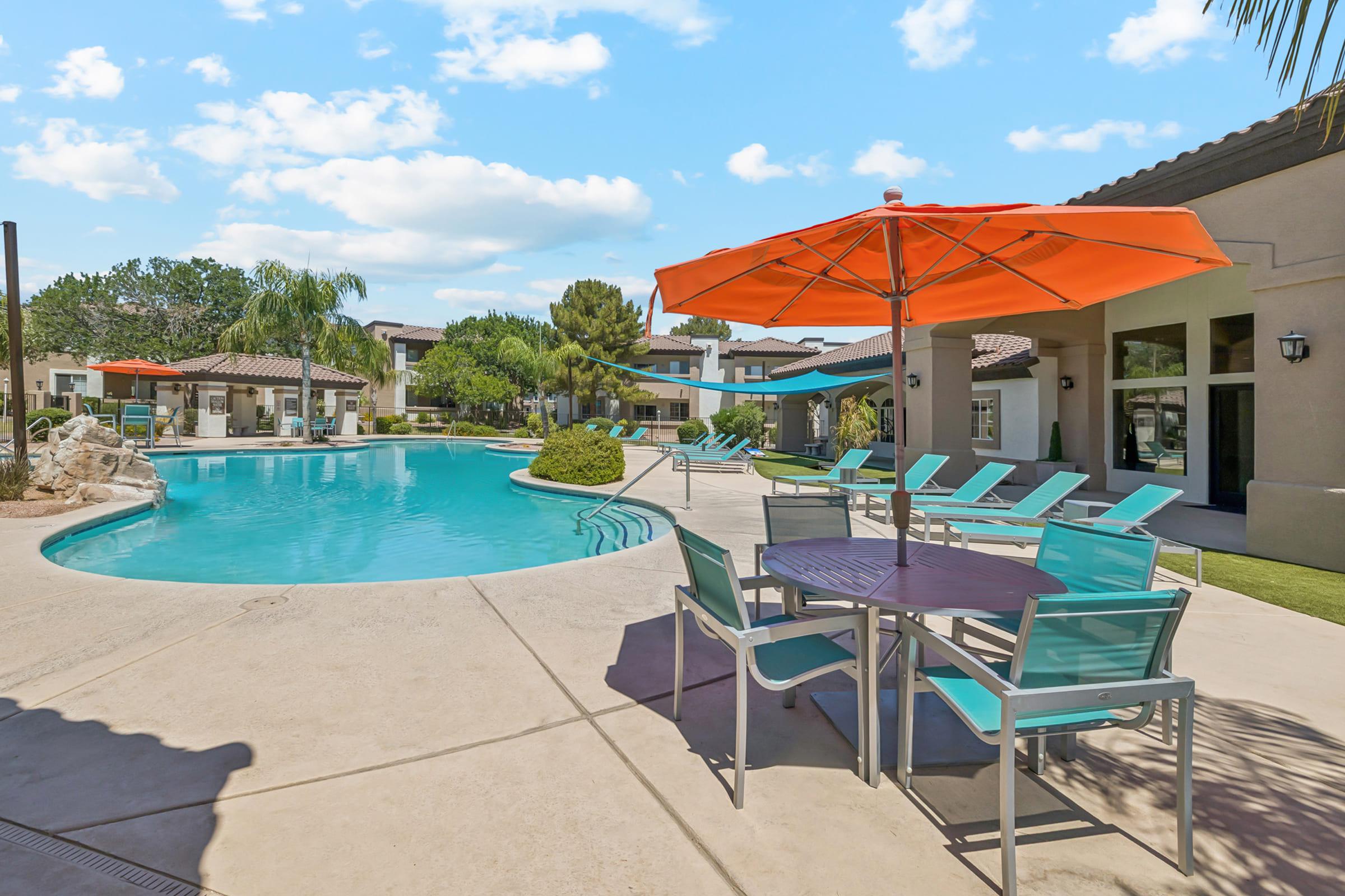 A sunny outdoor pool area featuring a turquoise swimming pool surrounded by lounge chairs and a table with an orange umbrella. Lush greenery and modern architecture are visible in the background. The sky is clear with fluffy clouds, creating a relaxing atmosphere.