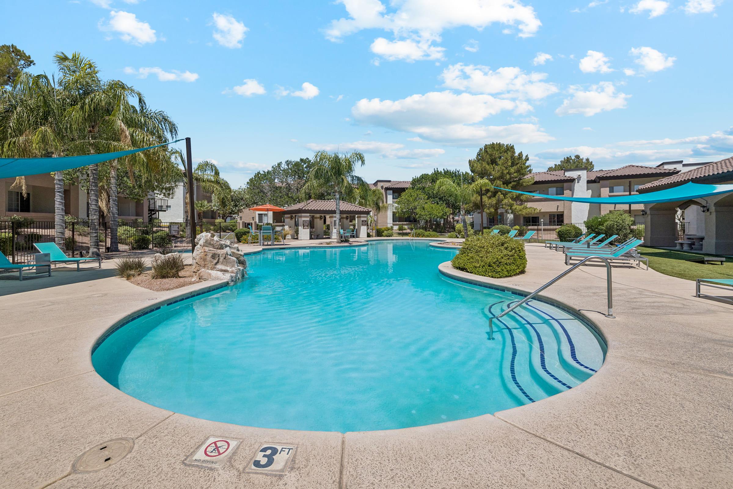 A serene swimming pool area featuring clear blue water, surrounded by palm trees and lounge chairs. There are shaded areas with canopies nearby, and landscaped greenery adds to the relaxing atmosphere. A sign indicates the shallow end of the pool is 3 feet deep.