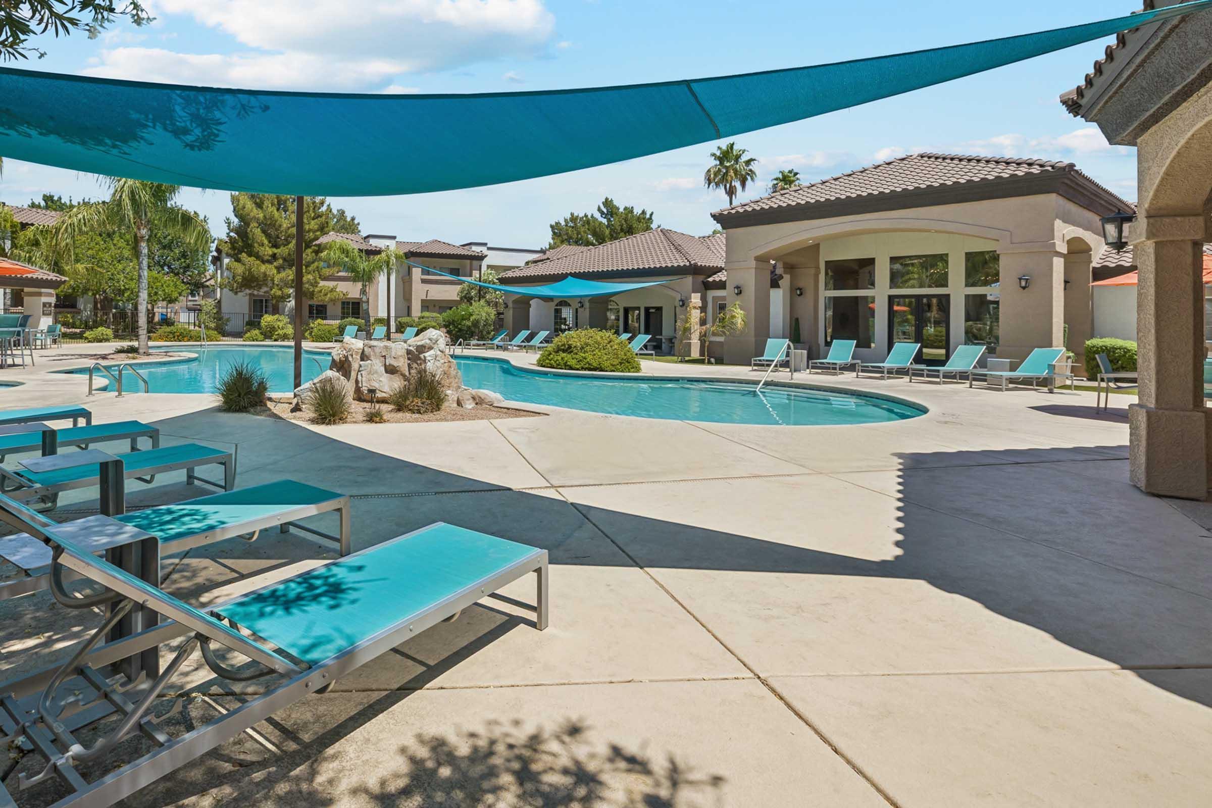 Pool area featuring a clear blue water pool surrounded by lounge chairs and shaded by a large canopy. There are palm trees and well-maintained landscaping, with nearby buildings visible in the background. The scene is bright and inviting, suggesting a relaxing outdoor space.
