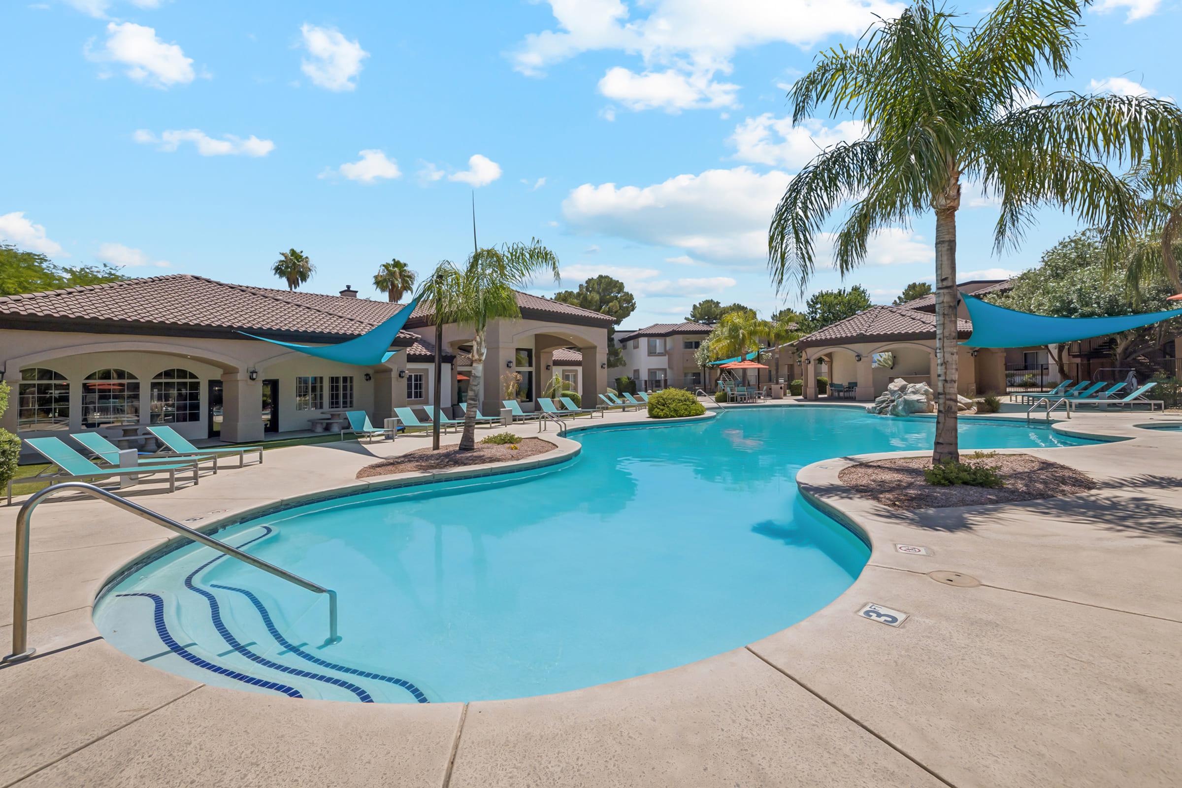 A tranquil swimming pool surrounded by palm trees and lounge chairs, with shaded areas created by blue canopies. The area features well-maintained landscaping and a relaxed atmosphere, ideal for leisure. In the background, there are residential buildings, adding to the inviting environment.