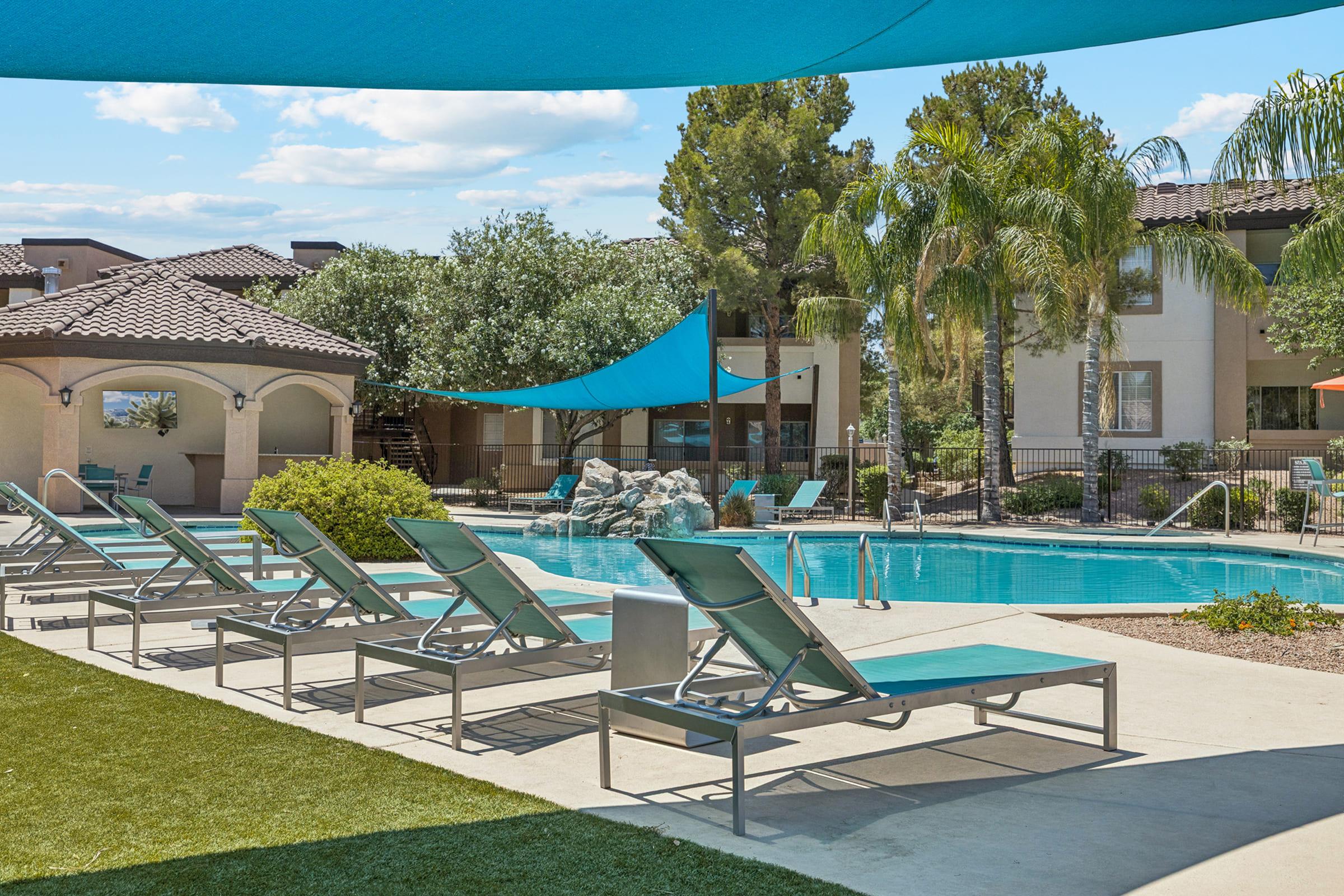 A serene pool area featuring lounge chairs arranged around a sparkling swimming pool. Shade sails provide cover from the sun, while palm trees and landscaped greenery enhance the relaxing atmosphere. The background showcases residential buildings and clear blue skies. Perfect for relaxation and leisure.