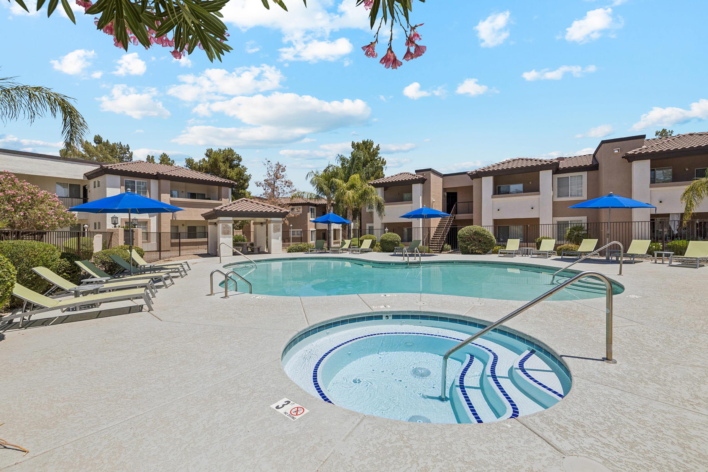 A swimming pool surrounded by lounge chairs and blue umbrellas, with a hot tub nearby. The pool area is landscaped with palm trees and flowers, and there are apartment buildings in the background under a partly cloudy sky.