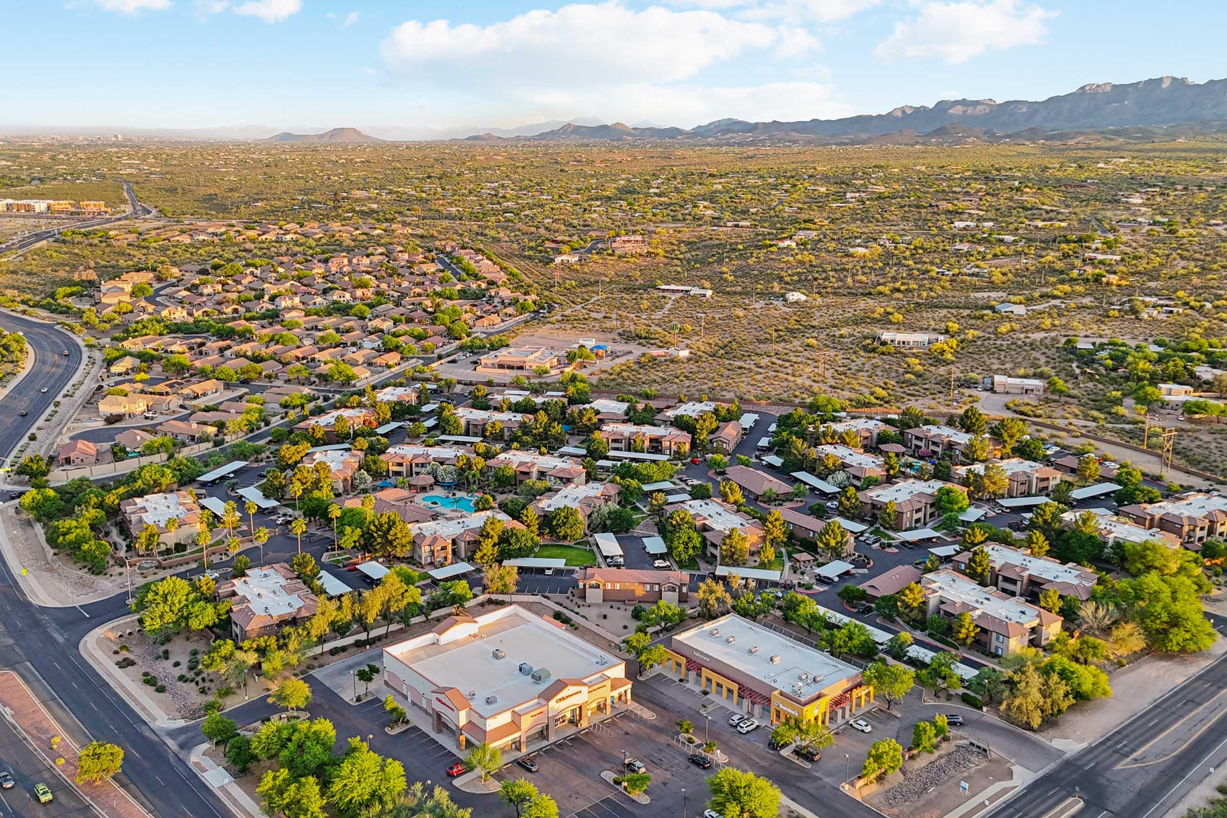 Aerial view of a suburban area featuring residential neighborhoods, pools, and nearby mountains under a blue sky with scattered clouds.