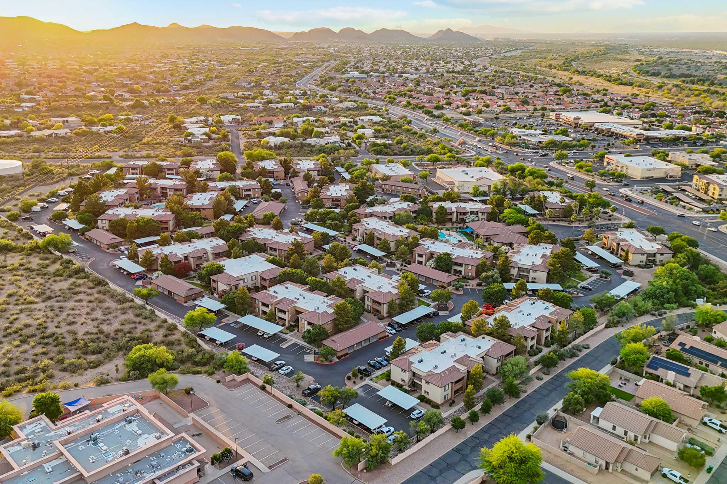 Aerial view of a residential neighborhood at sunset, featuring a cluster of buildings with flat roofs, surrounded by greenery and mountains in the background.
