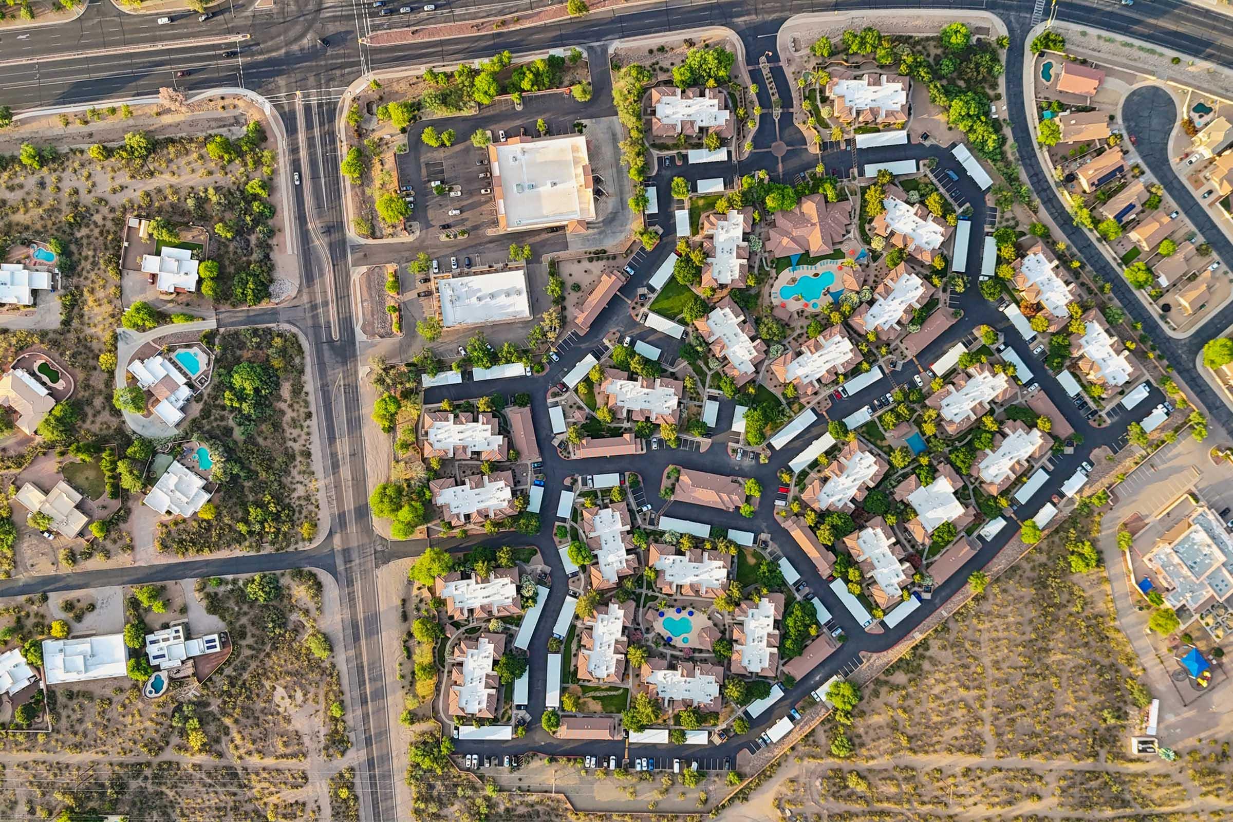 Aerial view of a residential community featuring several buildings arranged in a circular pattern, with green landscaping, swimming pools, and surrounding roads.
