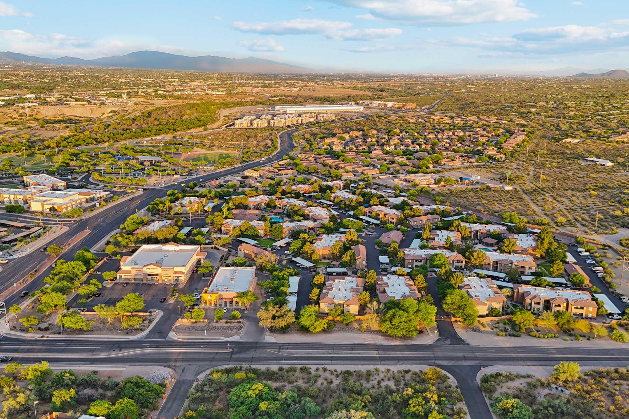 Aerial view of a suburban area with houses clustered among green trees, set against a backdrop of mountains and open land.