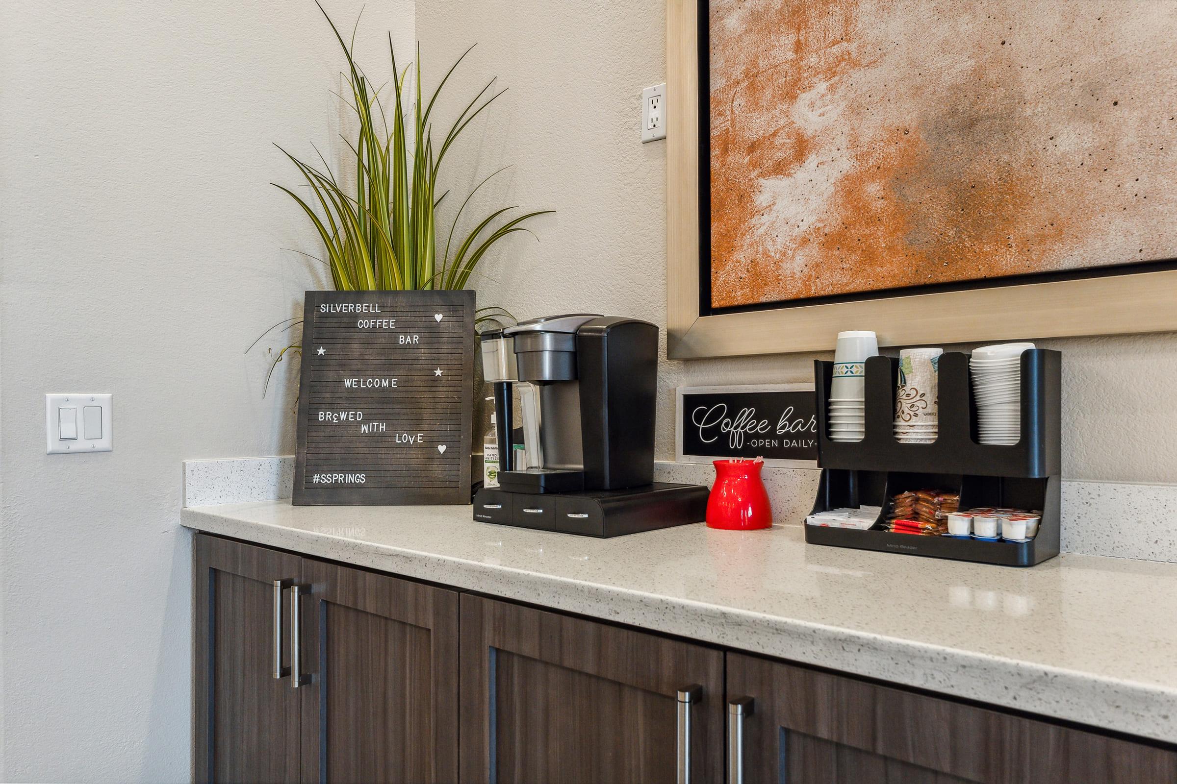 A coffee station featuring a coffee maker, a "Coffee Bar – Open Daily" sign, disposable cups, sweeteners, and a decorative plant on a countertop.
