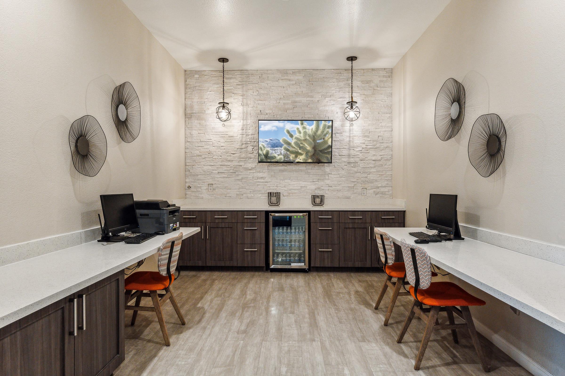 A modern shared workspace featuring two desks with computers, a small refrigerator, and decorative wall-mounted art.