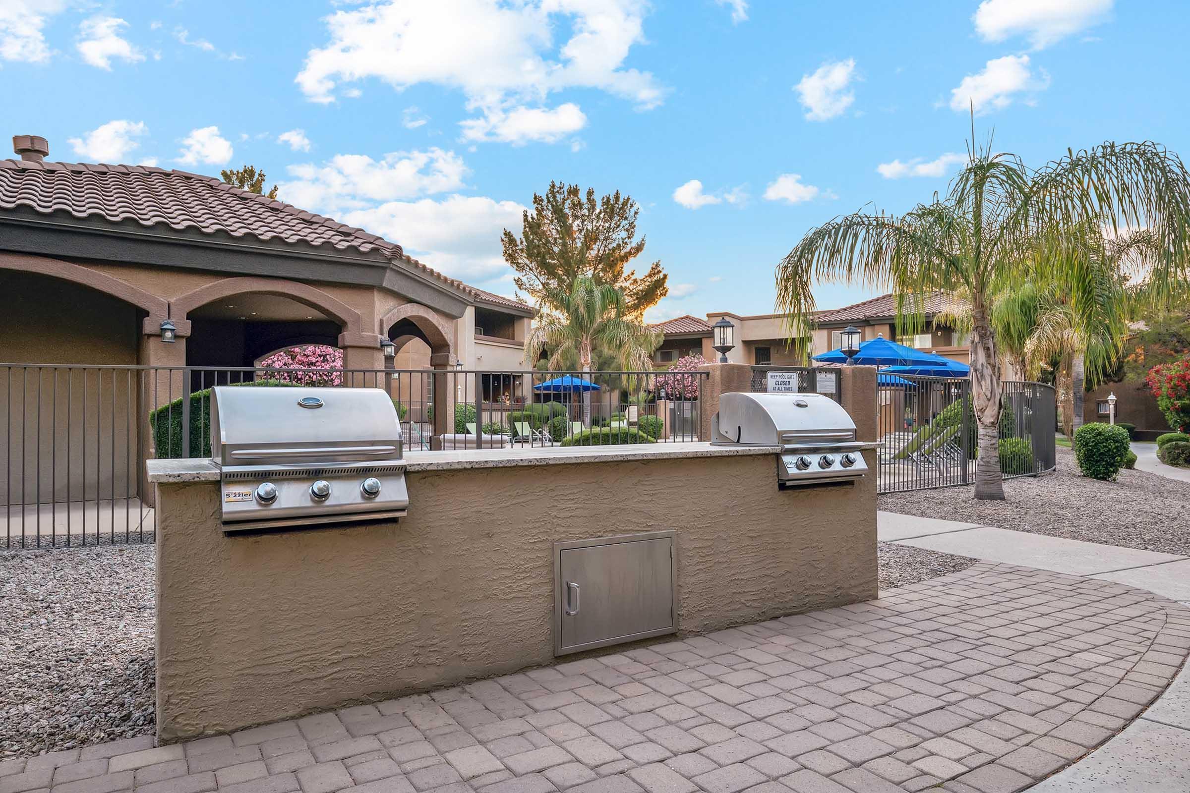 Outdoor grilling area featuring two stainless steel grills on a concrete counter, surrounded by landscaped greenery, palm trees, and outdoor seating.