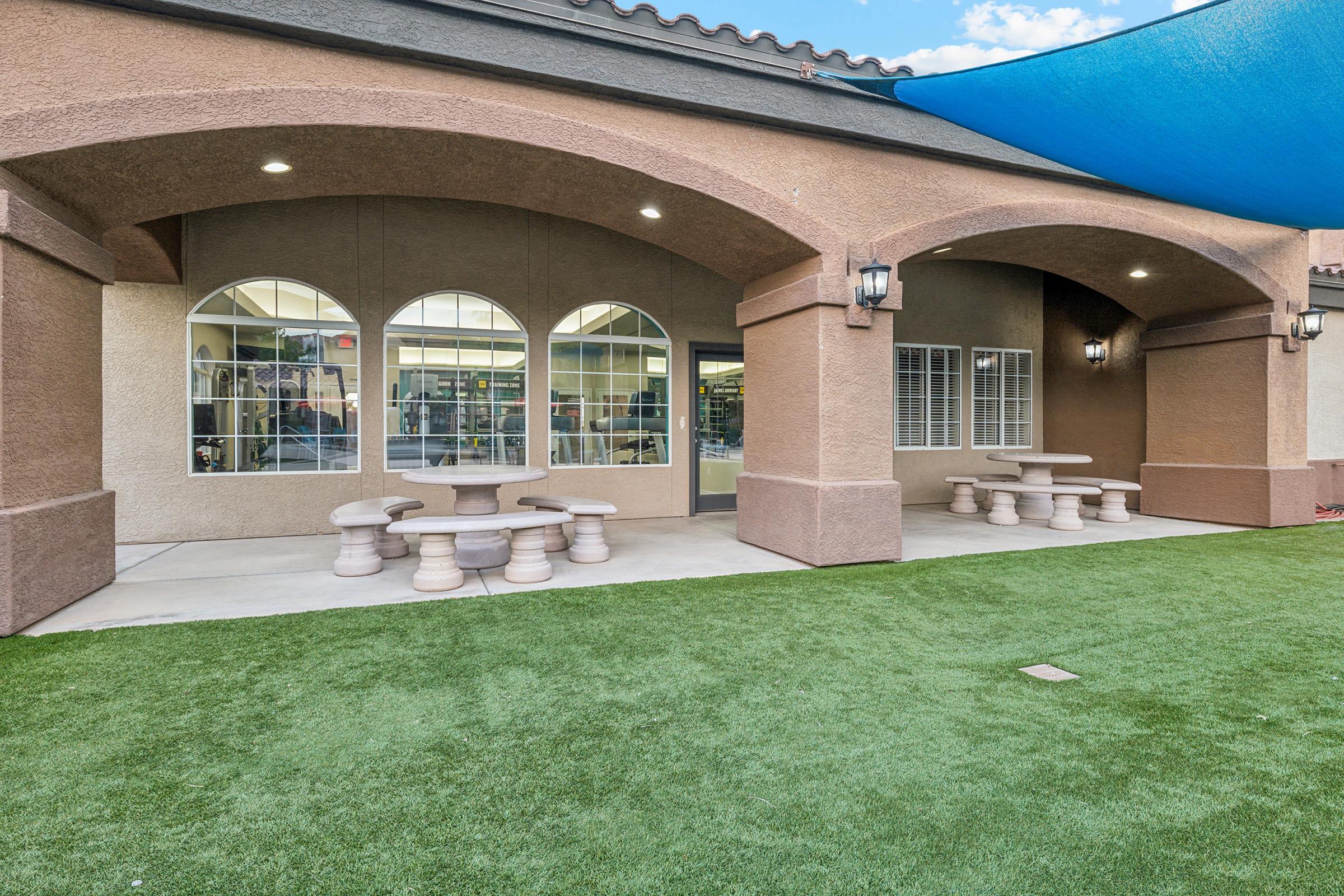 Outdoor seating area featuring a covered patio with arched windows, accompanied by round stone tables and benches on grass. T
