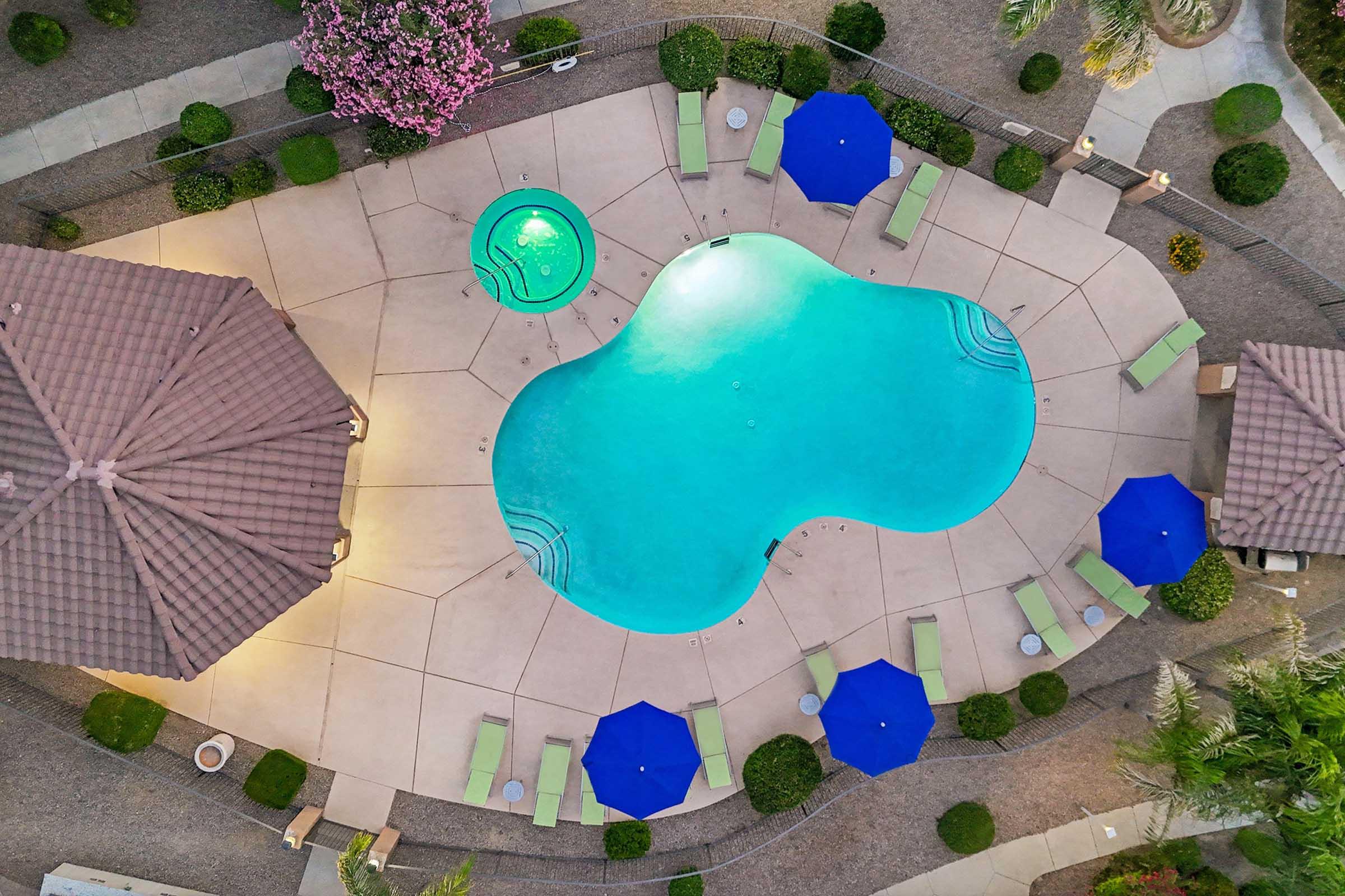 Aerial view of a resort-style swimming pool in a landscaped area. The pool has a unique shape, surrounded by green loungers and blue umbrellas.