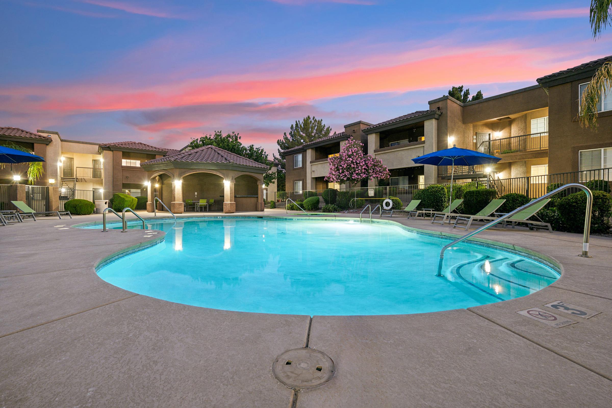 A serene swimming pool surrounded by landscaped greenery and chairs, with an apartment complex in the background.