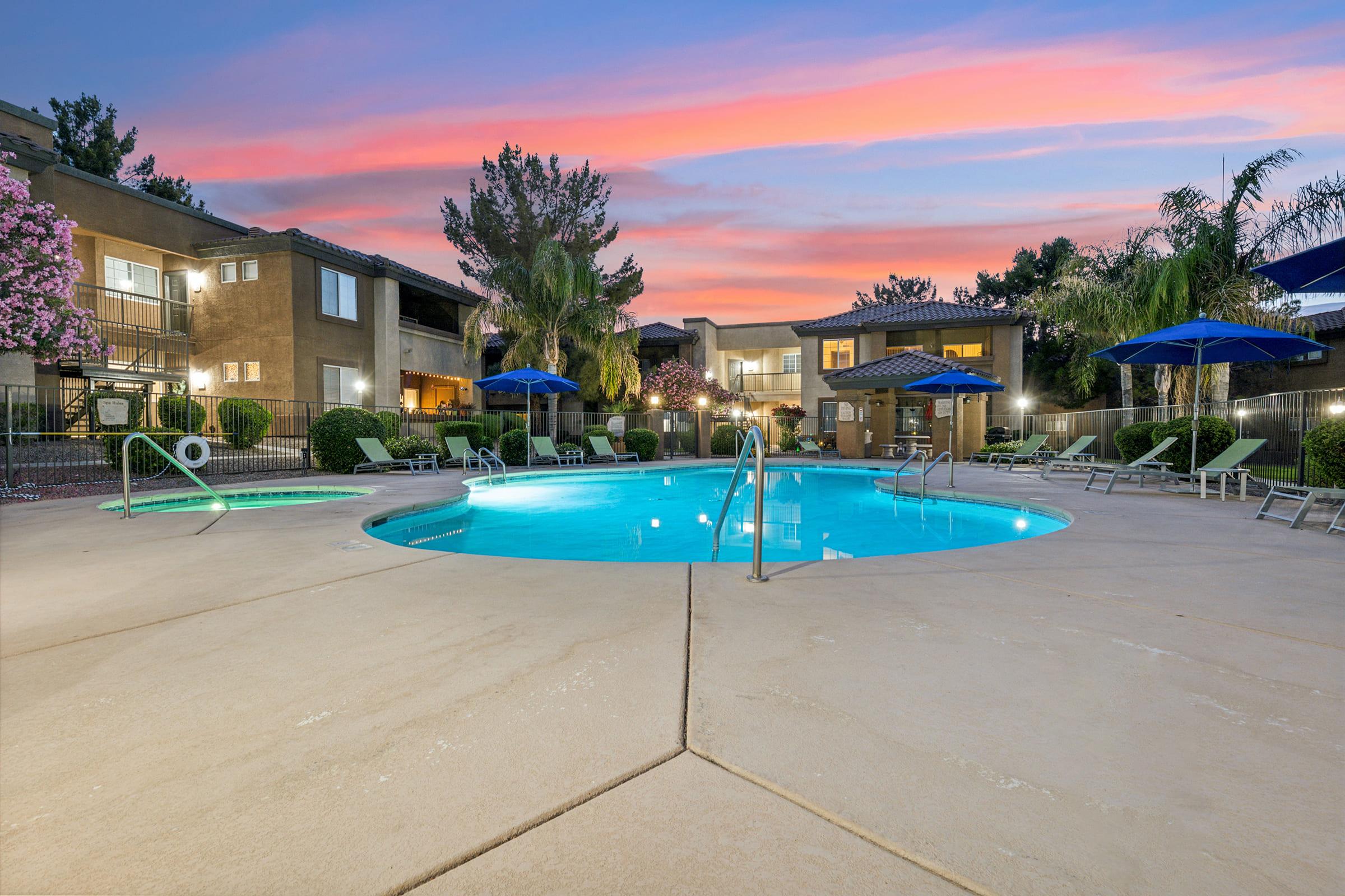 A serene swimming pool surrounded by lounge chairs and blue umbrellas, with palm trees and blooming flowers nearby.